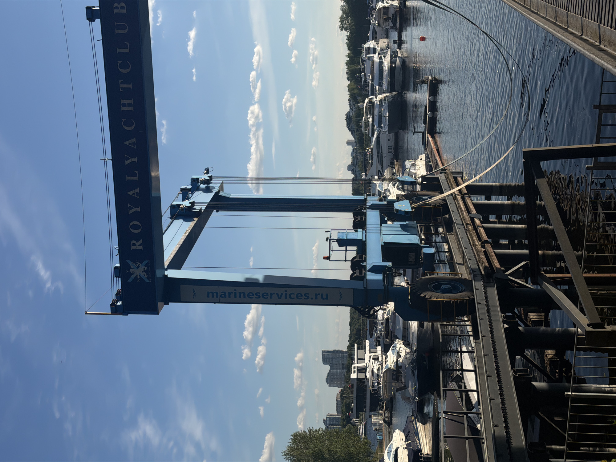 A blue marine travel lift with the text "ROYAL YACHT CLUB" visible on its upper beam stands on a pier extending over dark blue water. The travel lift has a large wheel on its right side. Several white and grey yachts are docked behind the pier in a marina. In the background, tall buildings are visible on the horizon, and a green tree is on the left. The sky is bright blue with scattered white clouds. The scene is set in the city of Khimki, Russia. There are no people clearly visible in the image.
