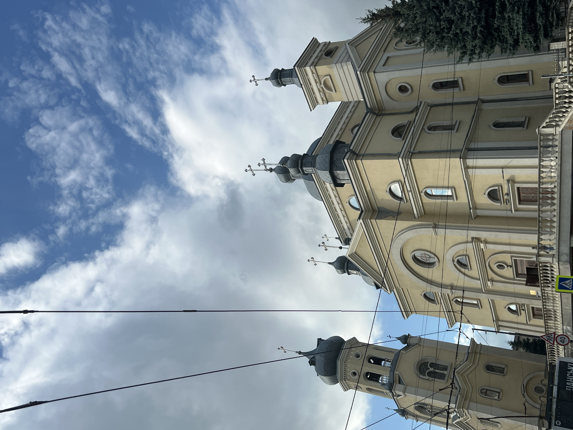 The image captures the Ternopil Theological Seminary building, also known as the Transfiguration Cathedral, in Ternopil, Ukraine. The architectural style is predominantly Baroque, with a prominent yellow facade adorned with white decorative elements. The building features multiple domes, topped with crosses, and a tall bell tower. The sky above is a mix of blue sky and dramatic grey clouds, with power lines crisscrossing the frame. In the foreground, a railing and steps suggest an elevated viewpoint or entrance. A few trees, including a dark green conifer on the right, are visible. Traffic signs, including a speed limit sign of 40 and a pedestrian crossing sign, are present near the bottom of the image, indicating a public street. One person is visible on the steps in front of the building.
