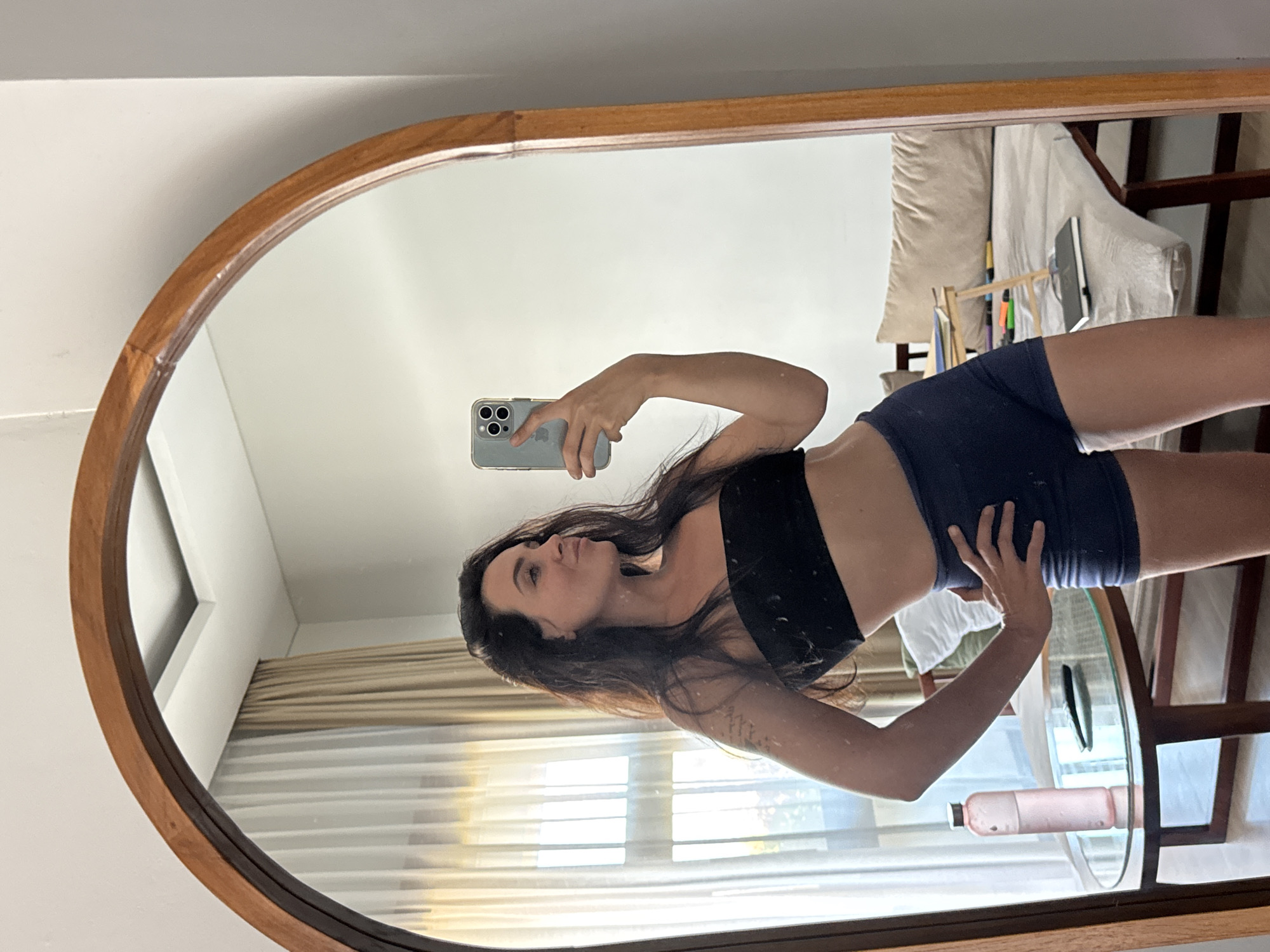 A woman with long dark hair is taking a mirror selfie in a room. She is wearing a black strapless top and navy blue athletic shorts. She is holding a light blue smartphone in her right hand, angled to capture her reflection. Her left hand is on her hip. In the mirror's reflection, behind her, a clear glass table holds a pink water bottle and a black object, possibly a remote. Further back, a bed is visible with a beige headboard and pillows. The room has sheer white and beige curtains covering a window. The overall setting suggests a hotel room or apartment in North Kuta, Indonesia.