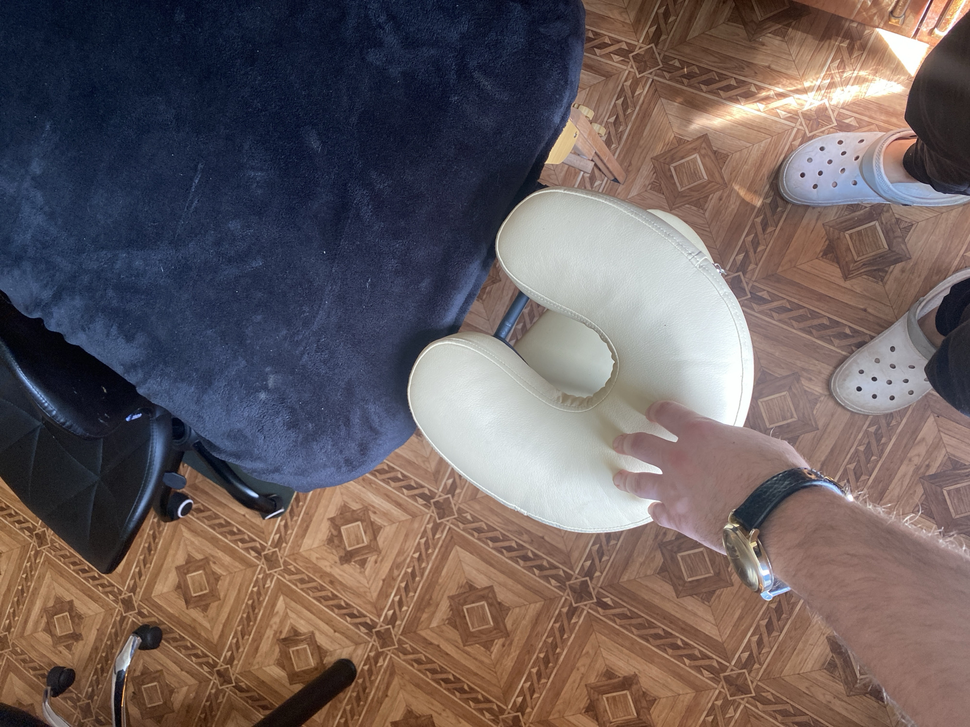 The image shows a person's arm with a watch on their wrist, reaching out to touch a pale yellow headrest. The headrest is part of a massage chair, which is covered with a dark blue plush fabric. The chair is on a wooden floor with a geometric pattern. In the background, a pair of white Crocs are visible, suggesting a casual or home setting. Sunlight is streaming in from the right, casting shadows and highlighting the floor's texture. The overall impression is a still moment, possibly before or after a massage. There is no visible text in the image.
