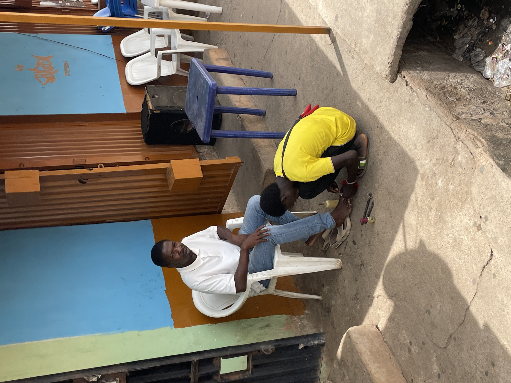 The image captures an outdoor scene in Jos, Nigeria, where two people interact. One man, wearing a white shirt and jeans, sits in a white plastic chair. Another person, wearing a yellow shirt, crouches near the seated man's feet, seemingly attending to them.
The setting includes a building with a blue and green wall, partially covered by wooden panels. Several white plastic chairs and a blue plastic table are positioned nearby. The ground is concrete, showing signs of wear and tear. A storm drain with visible debris is adjacent to the scene. A speaker is placed near the blue table and chairs.