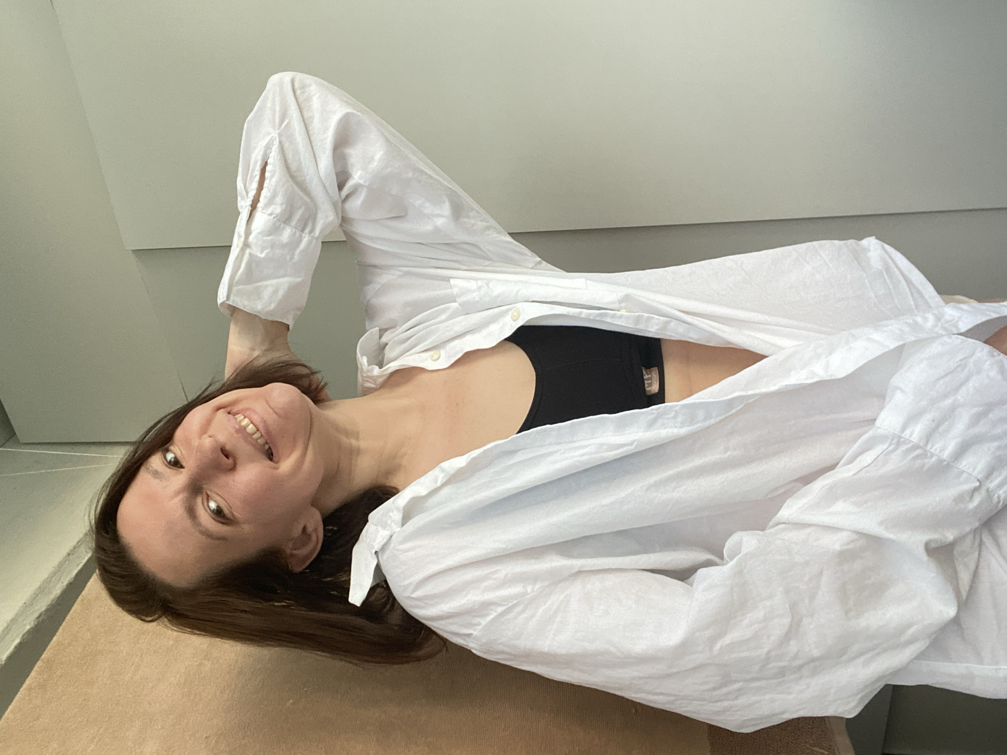 A woman with shoulder-length brown hair is lying on a beige surface, possibly a bed or similar furniture. She is wearing a black bra or tank top and a loose, open white shirt. She is smiling and looking towards the camera. The background consists of plain, light-colored walls, typical of an interior room. The location is not discernible from the image. The presence of a city such as Chaikovsky, Russia, cannot be determined.