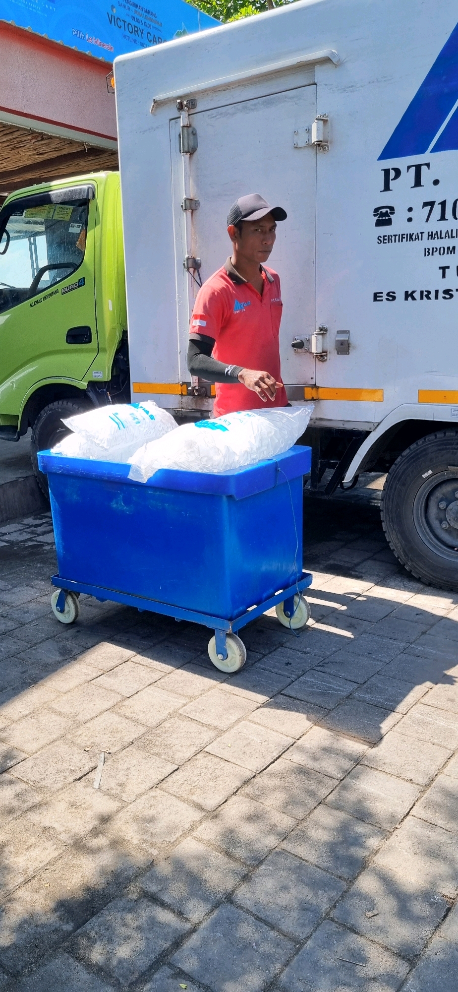 A man wearing a red shirt and a black baseball cap stands next to a white refrigerated truck and a blue cart filled with bags of ice. The truck has text on its side, including "PT." and a phone number. The cart has wheels and is positioned on a paved surface. In the background, a green truck is partially visible. The scene appears to be outdoors, possibly in a commercial or industrial area. There is signage visible on the white truck that reads "ES KRISTAL" and on a blue banner in the upper left corner that includes "VICTORY CARGO." The man is looking towards the right side of the image. Shadows from trees are cast on the paved ground.