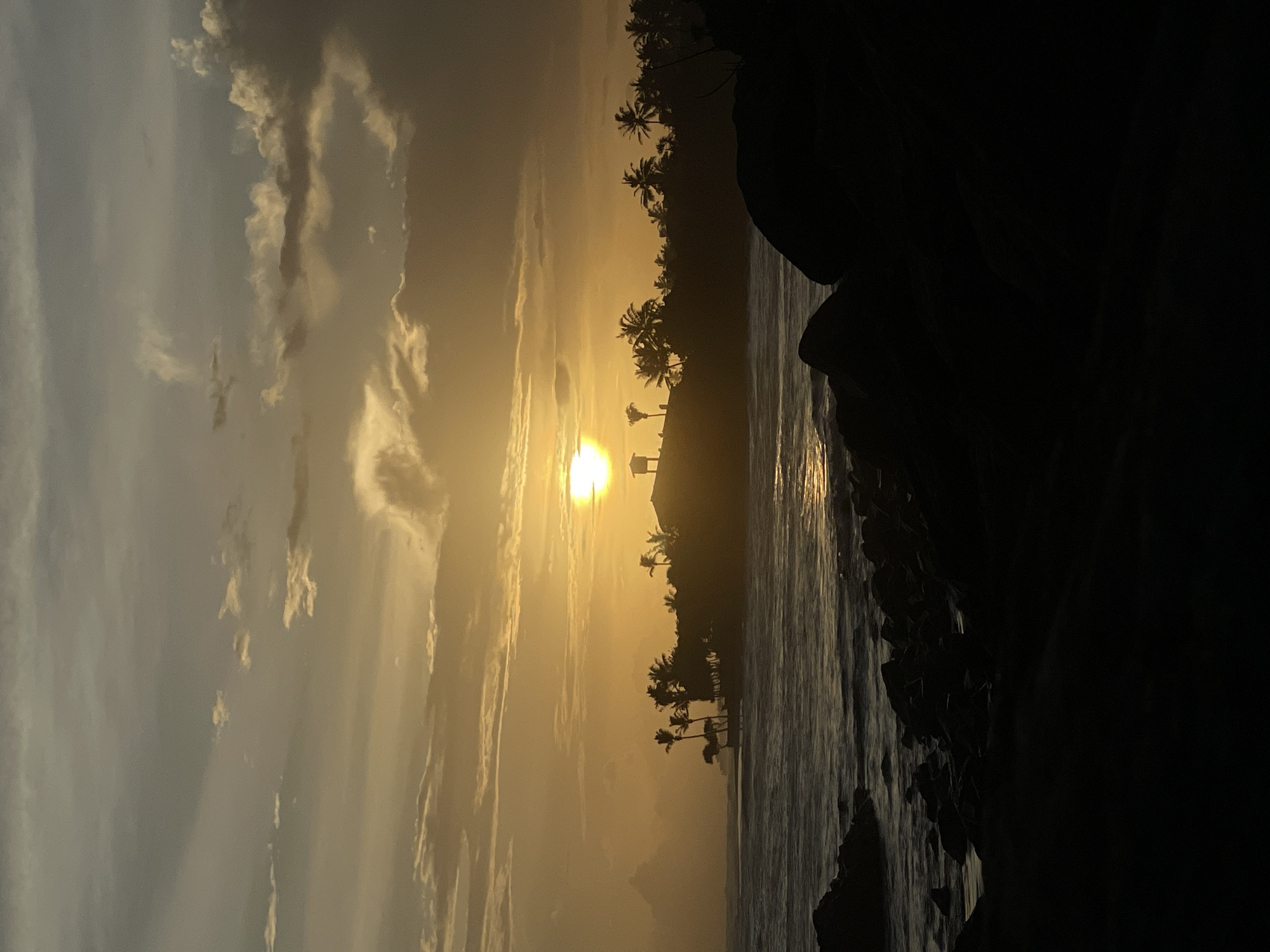 The image depicts a sunset over a body of water, likely the ocean, in Kathaluwa, Sri Lanka. The sun is positioned near the horizon, casting a golden glow across the sky and water. Silhouetted palm trees line the coast, and a structure, possibly a small building or shelter, is visible near the horizon. The foreground features dark rocks or boulders, creating a contrasting frame for the scene. The sky shows some cloud cover.