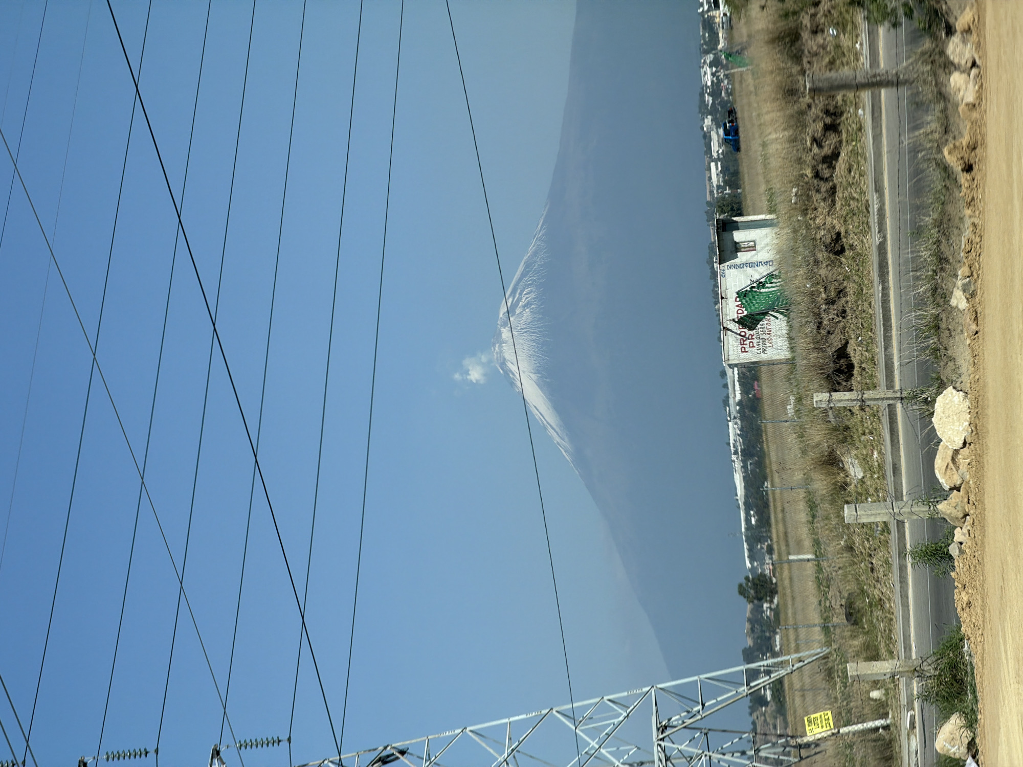 The image depicts a landscape in San Andrés Cholula, Mexico, featuring a view of Popocatépetl volcano under a blue sky. Power lines and a transmission tower are prominent in the foreground. Adjacent to a two-lane road is a grassy field, with a small building or structure bearing a faded sign in Spanish along the roadside. The general environment includes a mix of urban and rural elements. There are no people visible in the image.