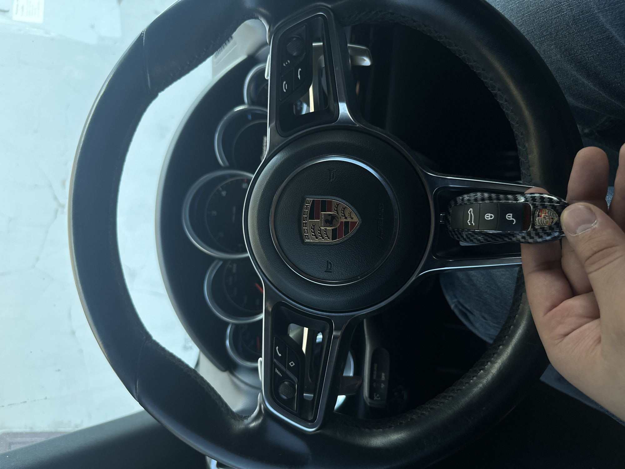 The image provides a close-up, high-angle view of the interior of a car, specifically the driver's side. The main subjects are a person's right hand holding a car key fob, and the car's steering wheel.
The steering wheel is black, leather-wrapped, and features metallic silver accents on its spokes. On the left spoke, there are control buttons for media and phone calls, including volume adjustment, track selection, and call answer/end functions. The center of the steering wheel prominently displays the colorful Porsche crest with the word "PORSCHE" inscribed below it. Faintly visible on the airbag cover is the word "AIRBAG."
The key fob, held by the person's hand, is encased in a cover with a distinctive black and white carbon fiber pattern. It features four silver buttons with symbols for unlocking the car, locking the car, opening the trunk, and a car locator/alarm. A small Porsche crest is also visible on the end of the key fob. The hand has visible fingernails.
Behind the steering wheel, parts of the car's instrument cluster are visible, showing dark circular gauges. A person's leg, clad in blue jeans, is partially seen in the lower right portion of the frame, behind the steering wheel. A stalk on the right side behind the steering wheel has faint text "RESUME" and "SET," likely related to cruise control.
The scene is brightly lit by diffused light entering from the left, suggesting it is daytime. The overall impression is a showcase of a Porsche car's interior and key, perhaps prior to or following a drive.