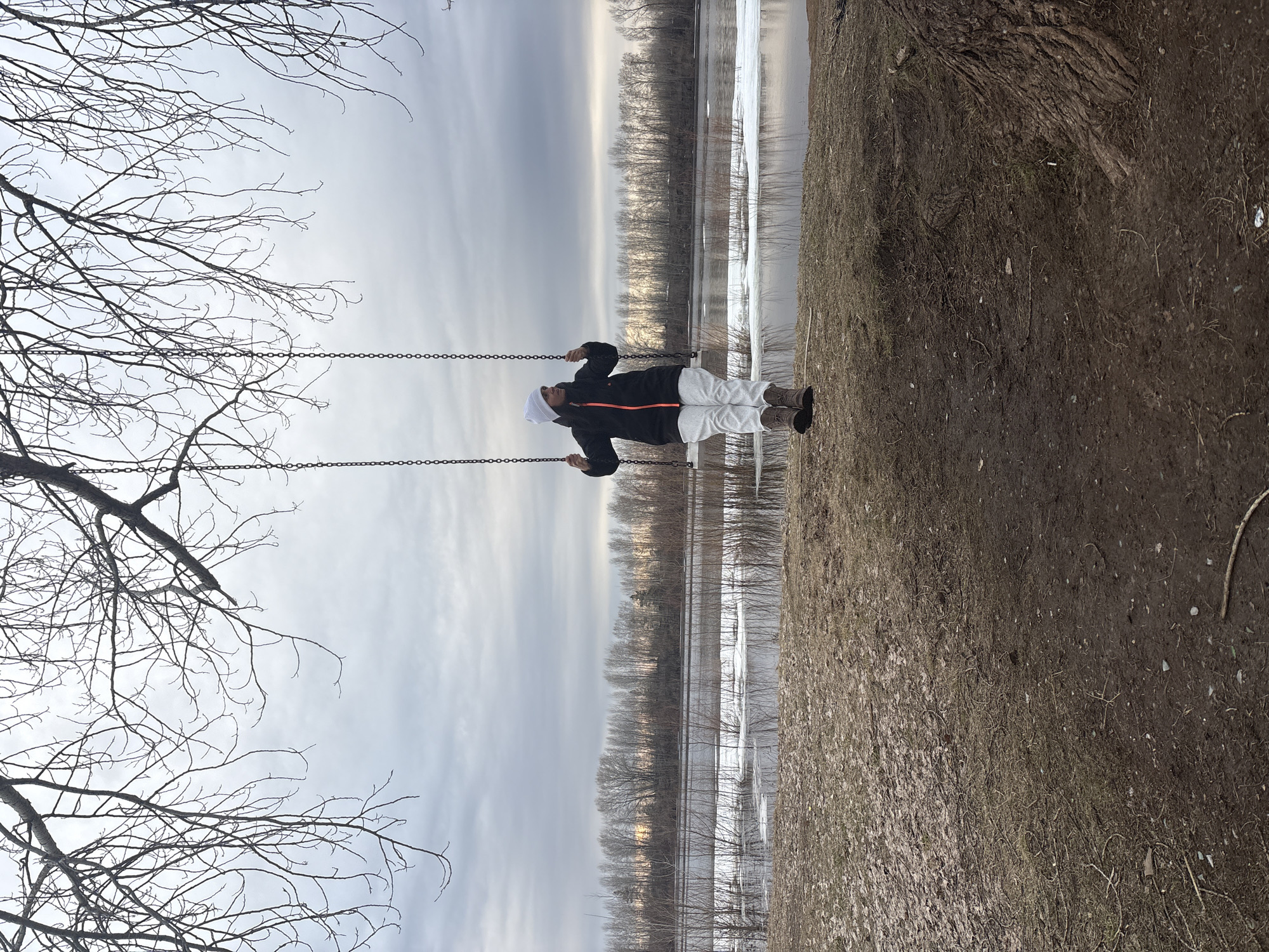 A person is sitting on a swing in a park. The location is Chaikovsky, Russia. The person is wearing a black jacket with an orange stripe, white pants, and brown boots. They also have a white head covering. The swing hangs from a tree with bare branches. Behind the swing is a body of water, possibly a river or lake, with some ice. Trees line the opposite bank. The ground is muddy and covered with dead leaves. The sky is overcast.
