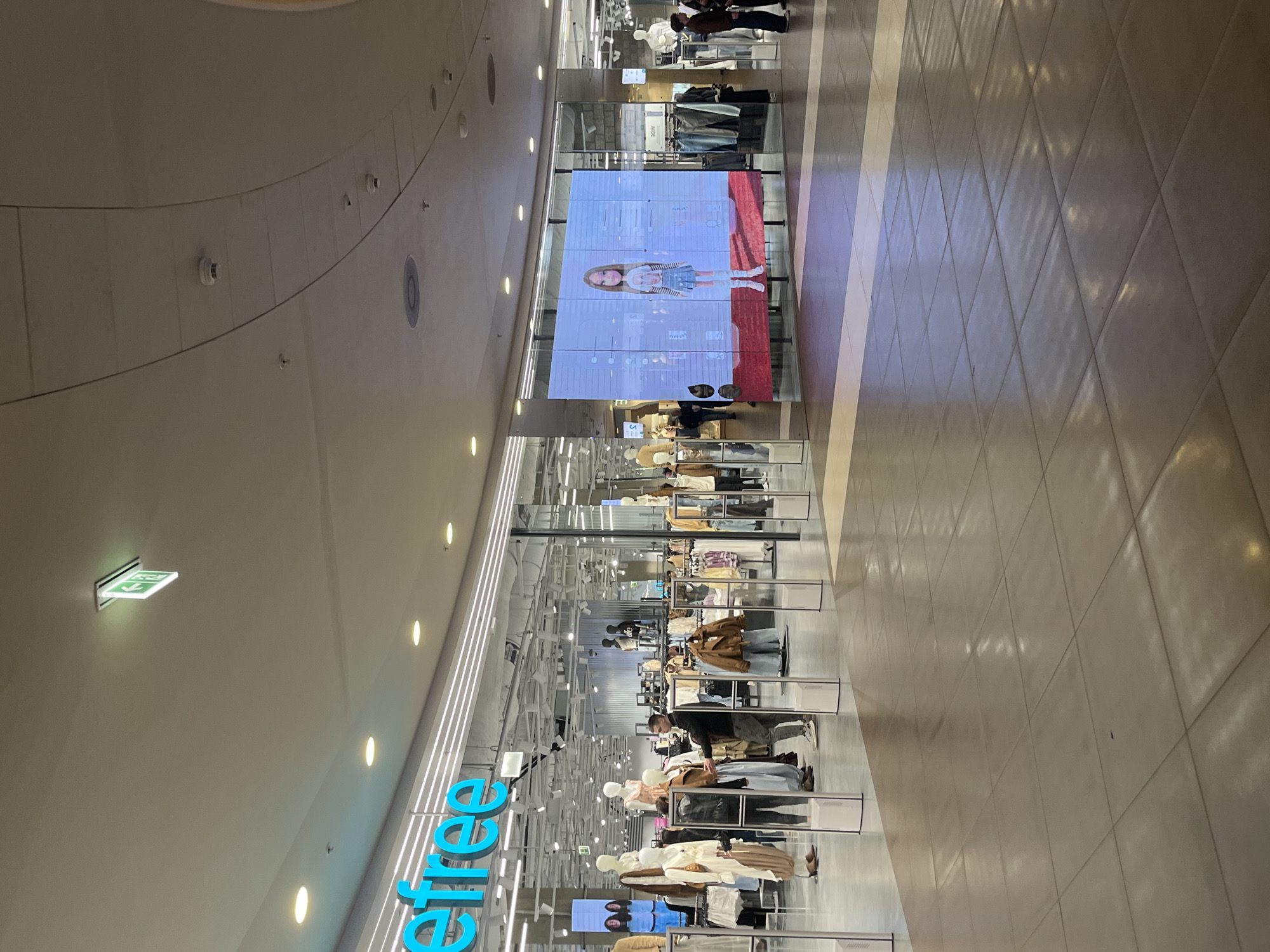 The image shows the interior of a large retail space, likely a clothing store, located in Saint Petersburg, Russia. The store, identified by the "elfree" sign, features mannequins displaying clothing items. A large LED screen displays a video of a child model. Customers are browsing the merchandise. The flooring is composed of large tiles, and the ceiling is high and curved.