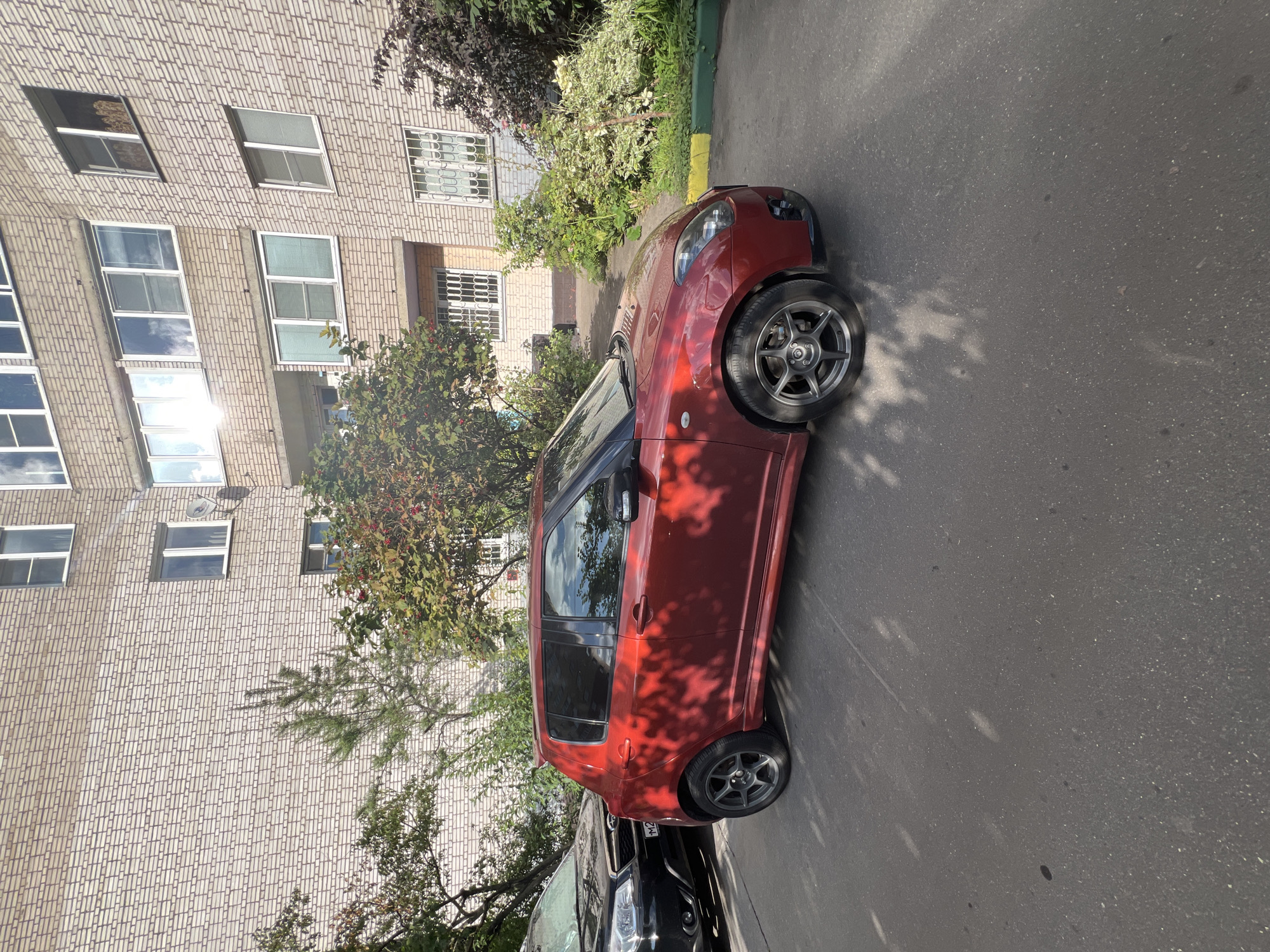 A red Suzuki Swift is parked on asphalt next to a black SUV in front of a brick apartment building. The building has multiple stories with many windows, some with white bars. There is greenery and trees between the cars and the building. The sun is shining, casting shadows from the trees onto the ground and the car. The scene appears to be in a residential area.