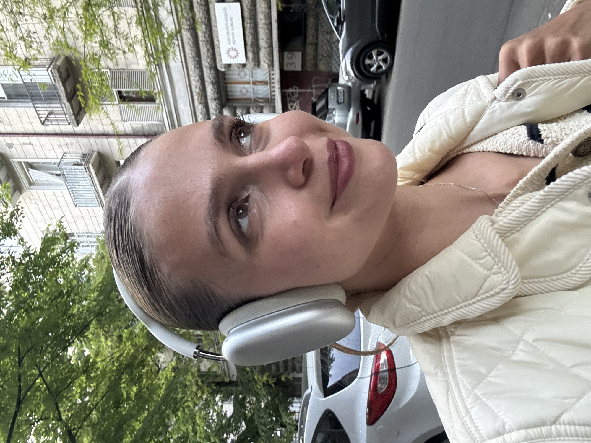 The image shows a young woman outdoors in what appears to be an urban setting in Tbilisi, Georgia. She is depicted from the chest up, with her head slightly tilted upwards and to her left (viewer's right). She has her hair pulled back, revealing her forehead, and features light brown or hazel eyes with a calm, slightly contemplative expression. She wears a rosy/mauve lip color.
She is wearing light-colored, over-ear headphones, identifiable as Apple AirPods Max, indicating she is listening to audio. Her attire includes a cream or off-white quilted jacket, and a light-colored, possibly knitted top with dark stripes underneath. A thin silver necklace is visible around her neck.
The background features multi-story buildings with windows and balconies, some equipped with shutters, consistent with typical urban architecture. Lush green foliage from trees is visible, suggesting a tree-lined street or a nearby park. Several parked cars can be seen on the street in the background. The natural, diffused lighting indicates it is daytime.
A sign on one of the buildings in the background clearly reads "LASHMI HOME DECOR" and the number "39," accompanied by a stylized lotus flower logo. This text provides a specific location detail within the city.