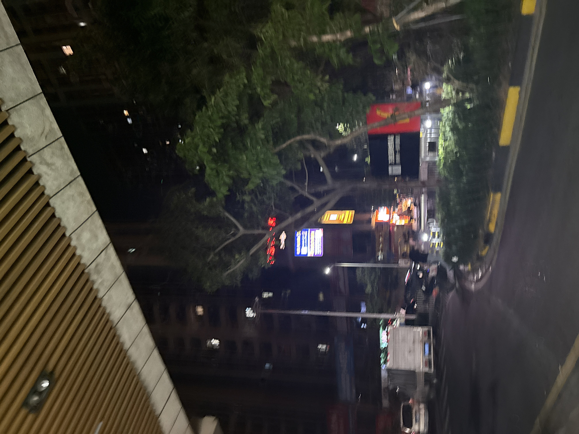 The image depicts a street scene at night in Haizhu District, China. A white truck is parked on the left side of the road. In the center of the image, a person is standing near a motorcycle. To the right, a red banner hangs vertically from a pole. The street is paved and has yellow and black curb markings. Buildings are visible in the background, with lights illuminating some windows. Trees and streetlights are also present in the scene. The overall impression is one of a typical urban environment after dark.
