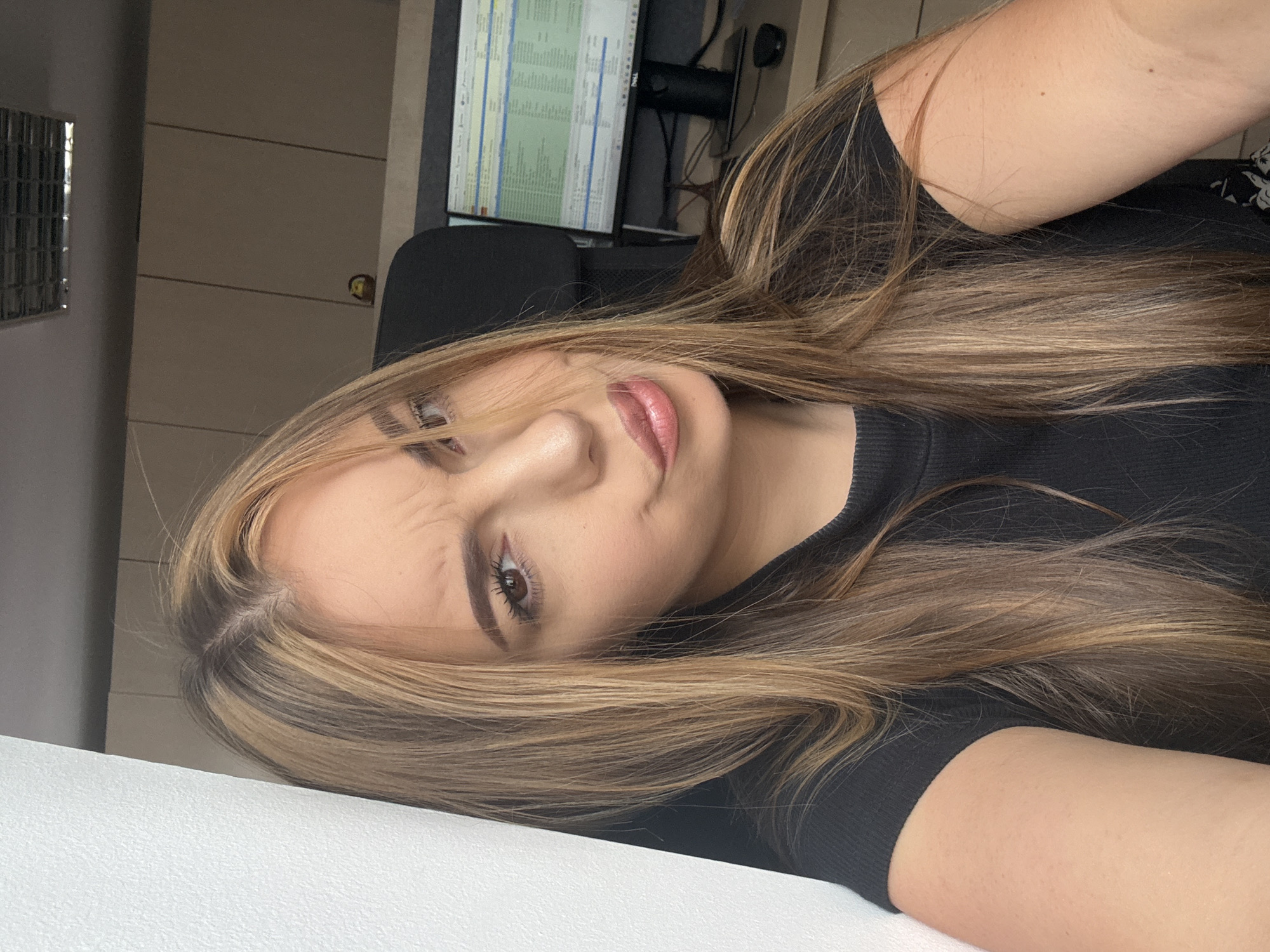 A young woman with blonde-streaked brown hair is taking a selfie in an office environment. She is wearing a black t-shirt and leaning against a white wall on the left. Her facial expression is a playful pout with raised eyebrows. In the background, a computer monitor displays a spreadsheet, and there are light brown cabinets mounted on the wall. The lighting suggests it's daytime. The scene is likely in Sofia, Bulgaria.