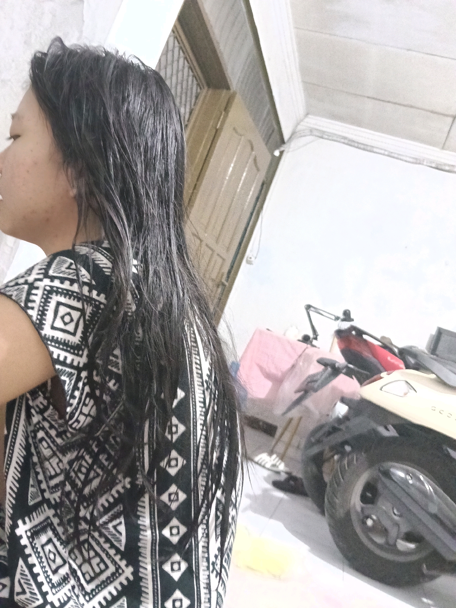 The image captures a medium shot of a young woman from the side, facing left, with her back to the camera. She has long, dark, wet hair that falls down her shoulders and back, and she is wearing a black and white patterned top. Her face is partially visible, showing her profile with closed eyes and her skin appears to have some blemishes.
The background suggests an outdoor or semi-outdoor setting, likely a residential area or alleyway. To the right, there are two motorcycles parked, one is a beige scooter and the other is partially visible with a red section. There are also some bags and other objects scattered in the background, including a pink bag. A wall with some wooden paneling is visible behind the woman. The overall lighting suggests it might be daytime, possibly overcast. The presence of motorcycles and the architectural style of the wall might indicate a location in a residential area in Jatimulya, Indonesia, known for its similar housing styles and common use of scooters as transportation.