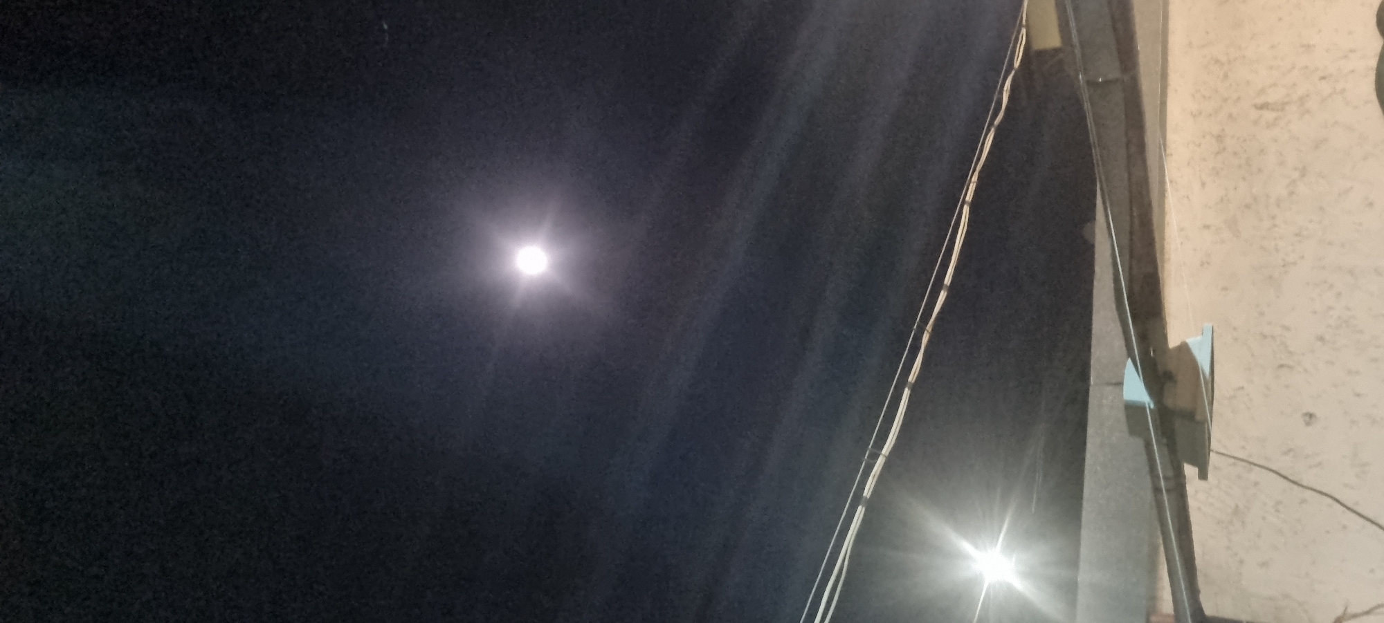 The image shows a dark night sky in an unknown city. A bright, circular object, potentially the moon, dominates the upper left portion of the frame. A set of wires runs diagonally across the image, intersecting with a structure attached to a light-colored wall on the right. A light source, possibly a street lamp, shines brightly near the base of the wall. No people or specific objects are identifiable beyond these elements.