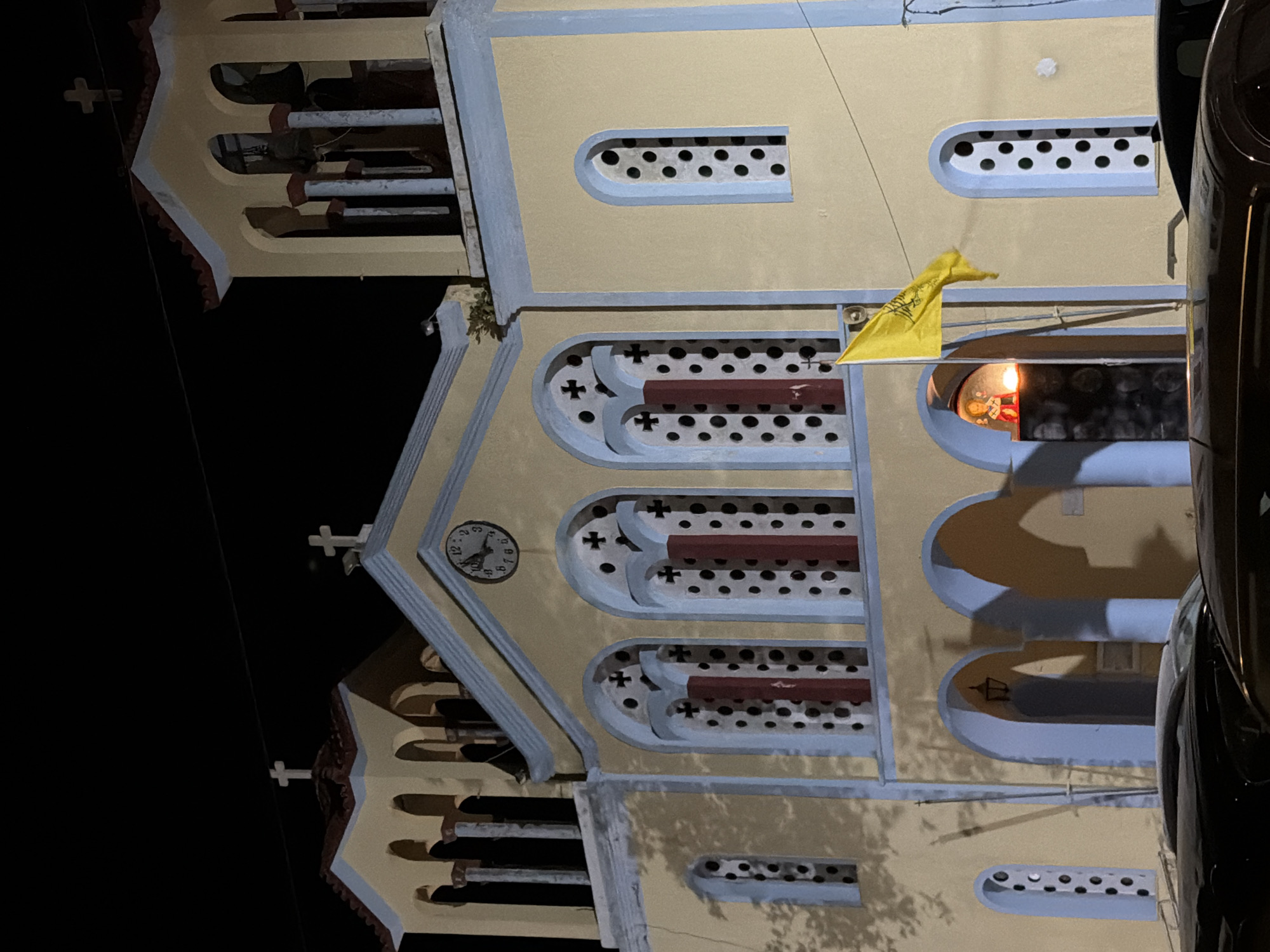 The image shows the exterior of a church at night, illuminated by artificial light. The church is painted in shades of yellow and light blue, with red accents. It has a clock on its facade, and a bell tower. There are several windows with decorative cross patterns and circular perforations. A yellow flag with an emblem is visible, attached to a pole. Within an arched entryway, a small statue of a religious figure can be seen, with a lit candle in front of it. The foreground is occupied by the tops of several dark-colored cars. The location is identified as the Municipal Unit of Alyzia, Greece.