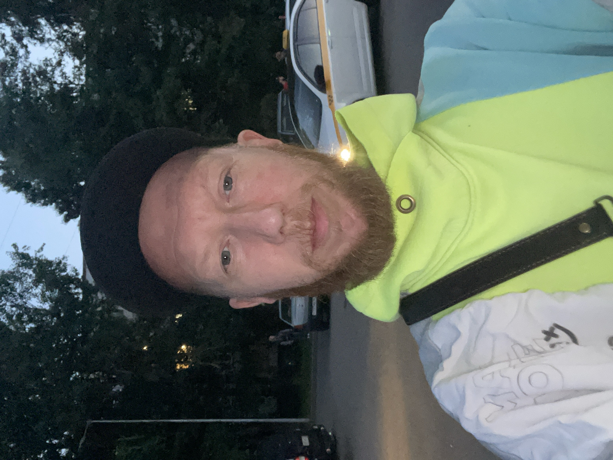 A man wearing a black beret and a brightly colored hoodie with sections of lime green and sky blue stands in a parking lot at dusk. He has a red beard and is looking directly at the camera. In the background, trees line the perimeter of the lot, and several cars are parked. A white taxi with a yellow stripe on its side is visible to the right of the man, with its headlights on. The lighting suggests the image was taken in an urban setting, possibly in Saint Petersburg, Russia, given the architectural style of the building in the background and the presence of a taxi that is commonly seen in Russian cities. The overall atmosphere is casual, with the man appearing to be taking a selfie.