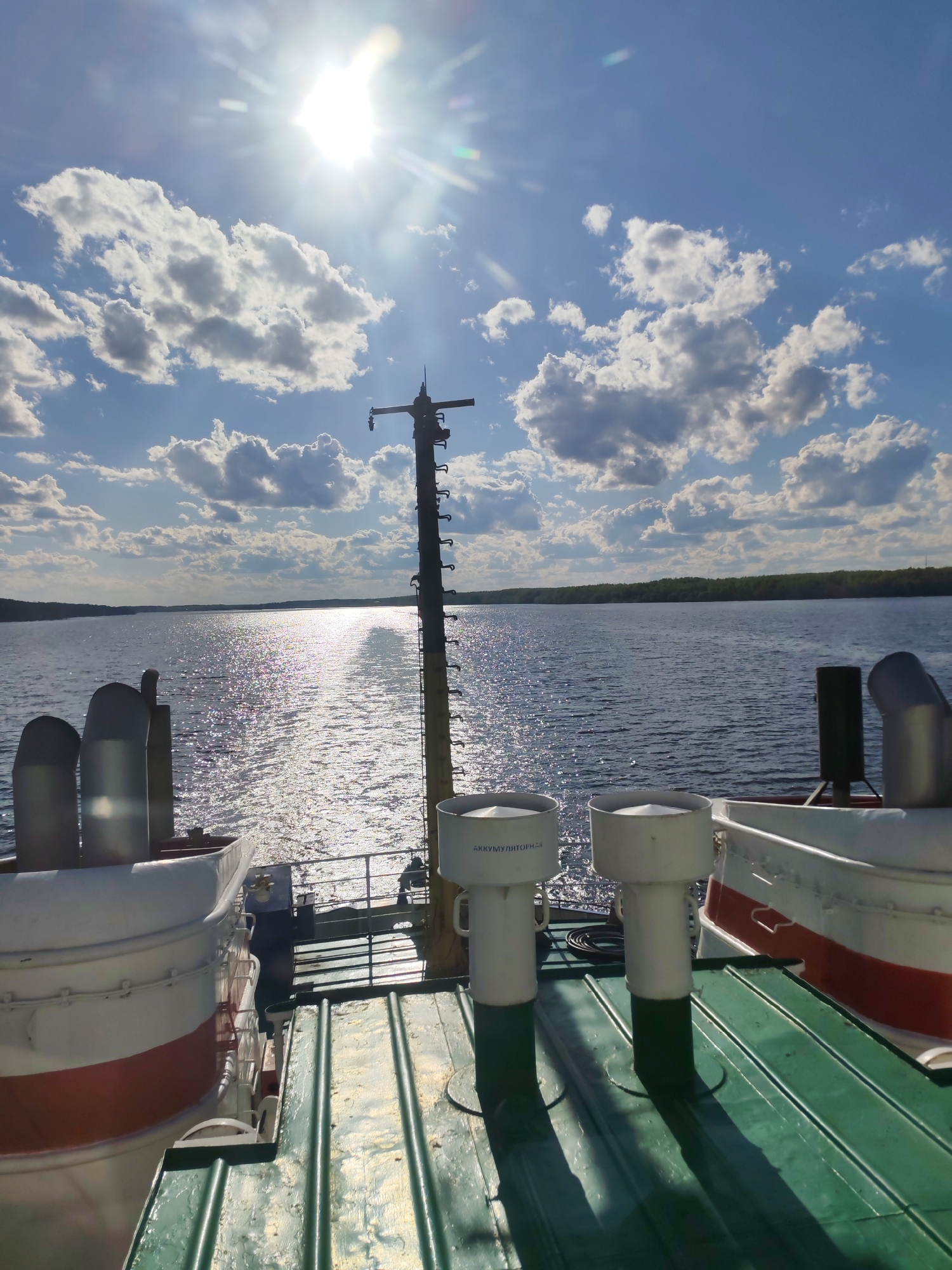 The image depicts a sunny, partly cloudy day on the Volga River in Myshkin, Russia. The view is from the deck of a vessel, looking towards the river. The sun is high in the sky, causing a bright reflection on the water's surface. The river is bordered by tree-lined shores in the distance. The deck of the ship is green and metallic, with various white and red ship components visible, including vents. A tall, metal mast with several attachments stands in the center of the view. The sky is blue with puffy white clouds.