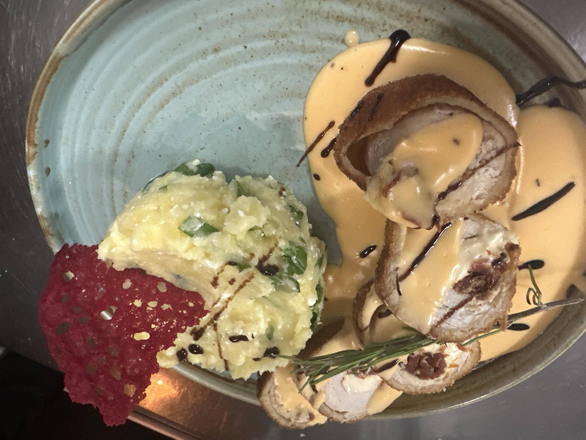 A top-down shot depicts a plate of food, potentially from a restaurant in Razlog, Bulgaria, given the context of the query. The plate, a light blue-green with brown edges, holds a portion of mashed potatoes with visible green herbs and a red crispy element. Sliced, breaded meat rolls are arranged alongside, coated in a pale orange sauce with dark brown drizzles. A sprig of green herb, possibly rosemary, rests on the meat. The plate sits on a metallic surface.