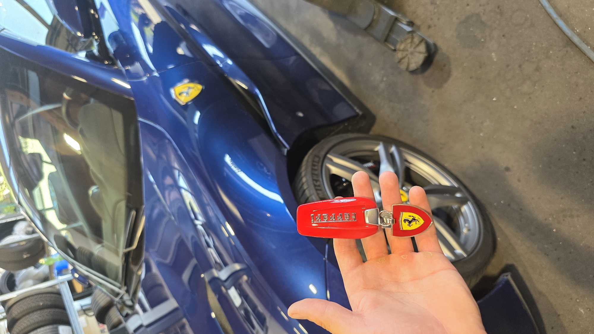 A person's hand holds a red Ferrari car key fob with the Ferrari logo at the bottom. The key fob is in focus and positioned in the foreground. Behind the key fob, a dark blue Ferrari car is visible. The car is partially obscured by the key fob, but its hood, fender, and a wheel are clearly discernible. The Ferrari emblem is also visible on the car's fender. The background shows a concrete floor, a tire rack filled with tires, and a hydraulic car jack. The lighting suggests an indoor setting, possibly a garage or workshop. The overall scene depicts a close-up view of a Ferrari car key and the car it belongs to.