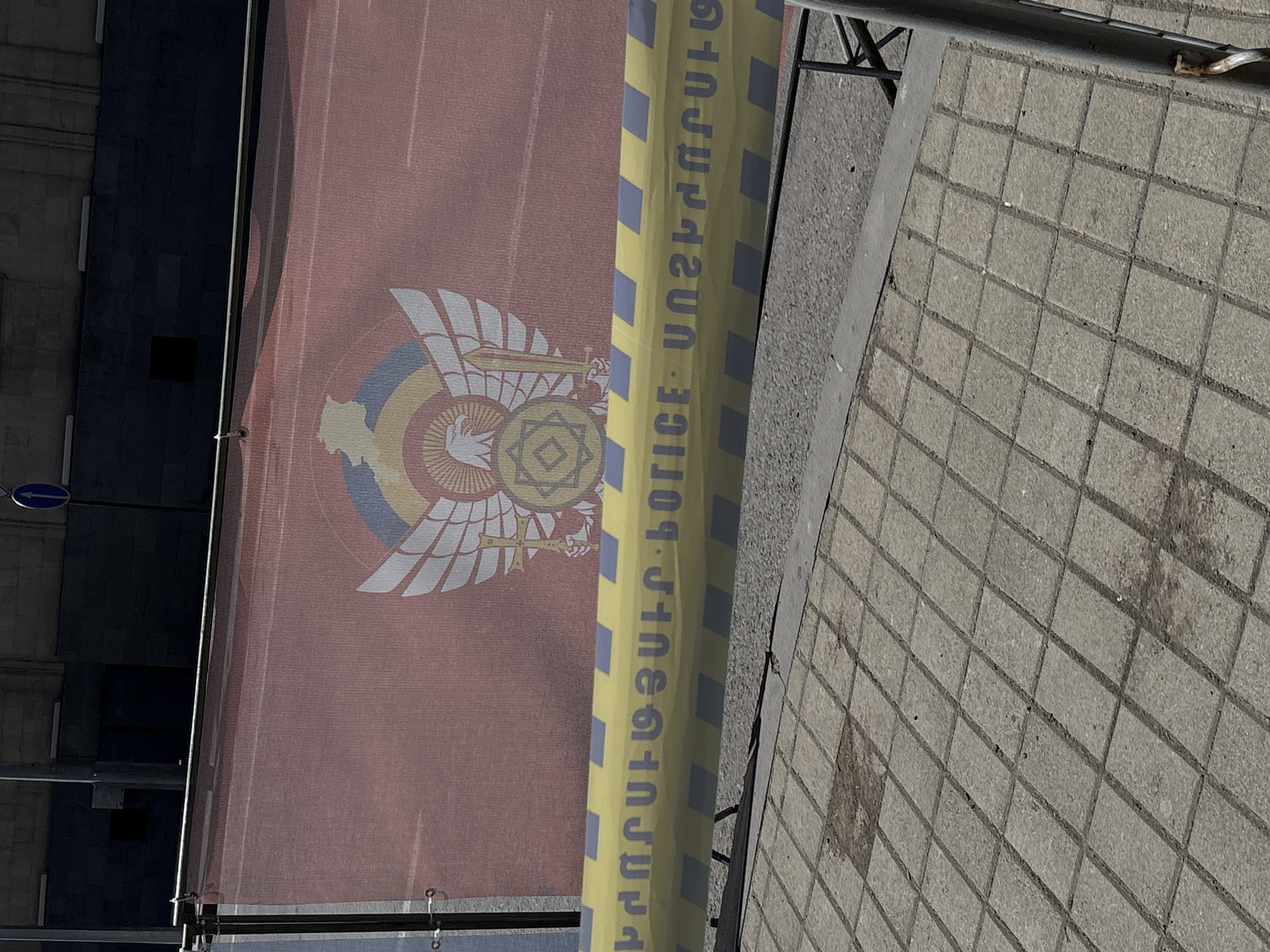 A ground-level, eye-level shot shows a banner featuring a stylized eagle with a cross design on a reddish background, obscured by a yellow police tape with Armenian text. The tape runs vertically. The pavement in the foreground features a diamond grid pattern. The banner is located in Yerevan, Armenia, based on the text on the tape.