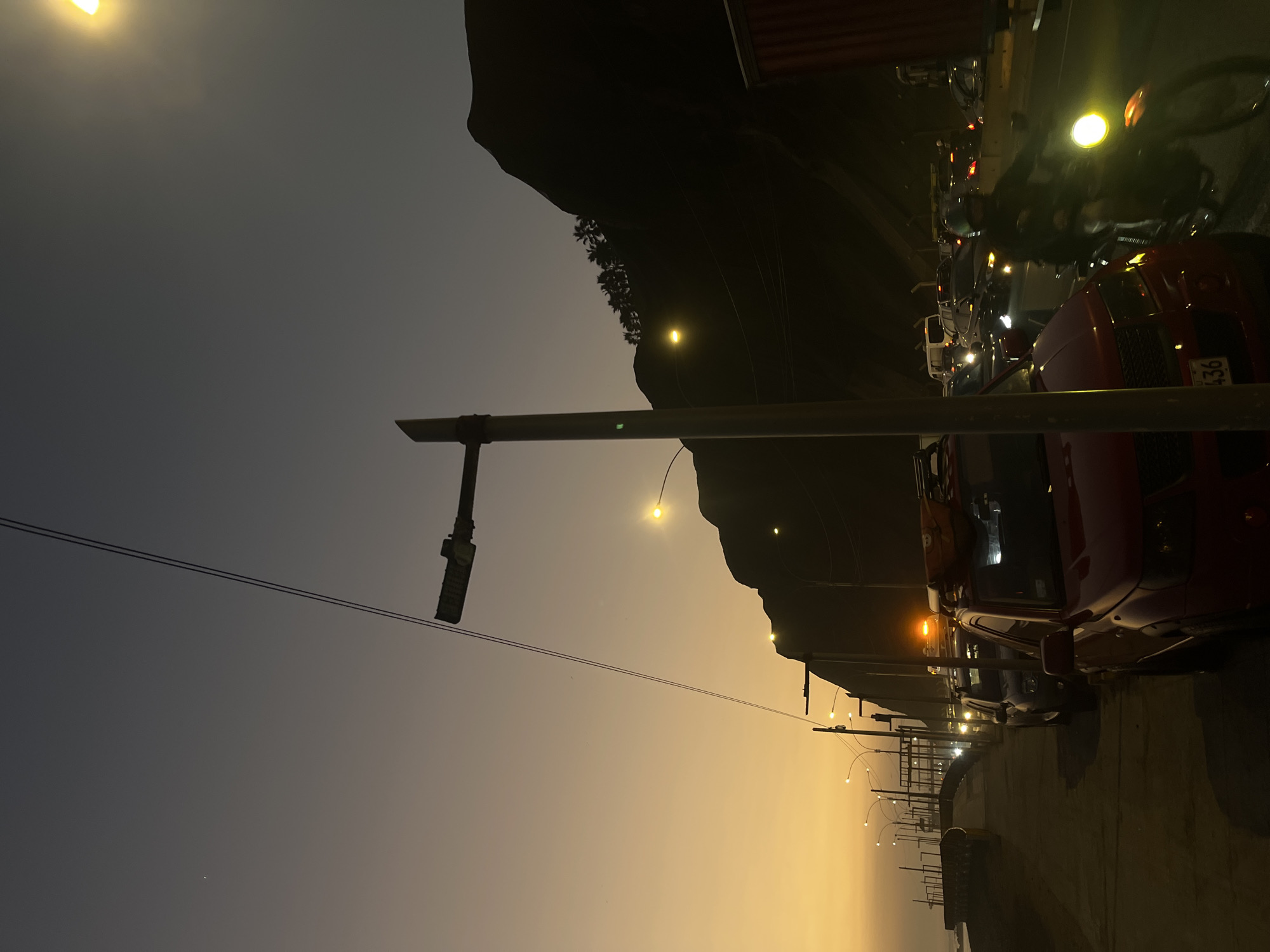 The image depicts a coastal road in Miraflores, Peru, at dusk. The sky transitions from a warm orange near the horizon to a deep gray overhead, with a faint star visible. Silhouetted against the sky are a mountain range, likely the Costa Verde cliffside, topped with palm trees, and several streetlights illuminating the scene. A power line runs diagonally across the frame.
In the foreground, a red SUV is parked, with a portion of its license plate visible, reading "436". Next to it, a motorcycle with its headlight on is being ridden by a person wearing a helmet. In the background, there is heavy traffic, with cars and trucks lining the road, their headlights and taillights creating streaks of light. Some vehicles have roof racks, suggesting they are either equipped for travel or recreational activities. The road appears to be a coastal highway, with a glimpse of the ocean to the left and a pedestrian walkway. The overall atmosphere is one of urban activity at the end of the day, with the natural landscape providing a dramatic backdrop.