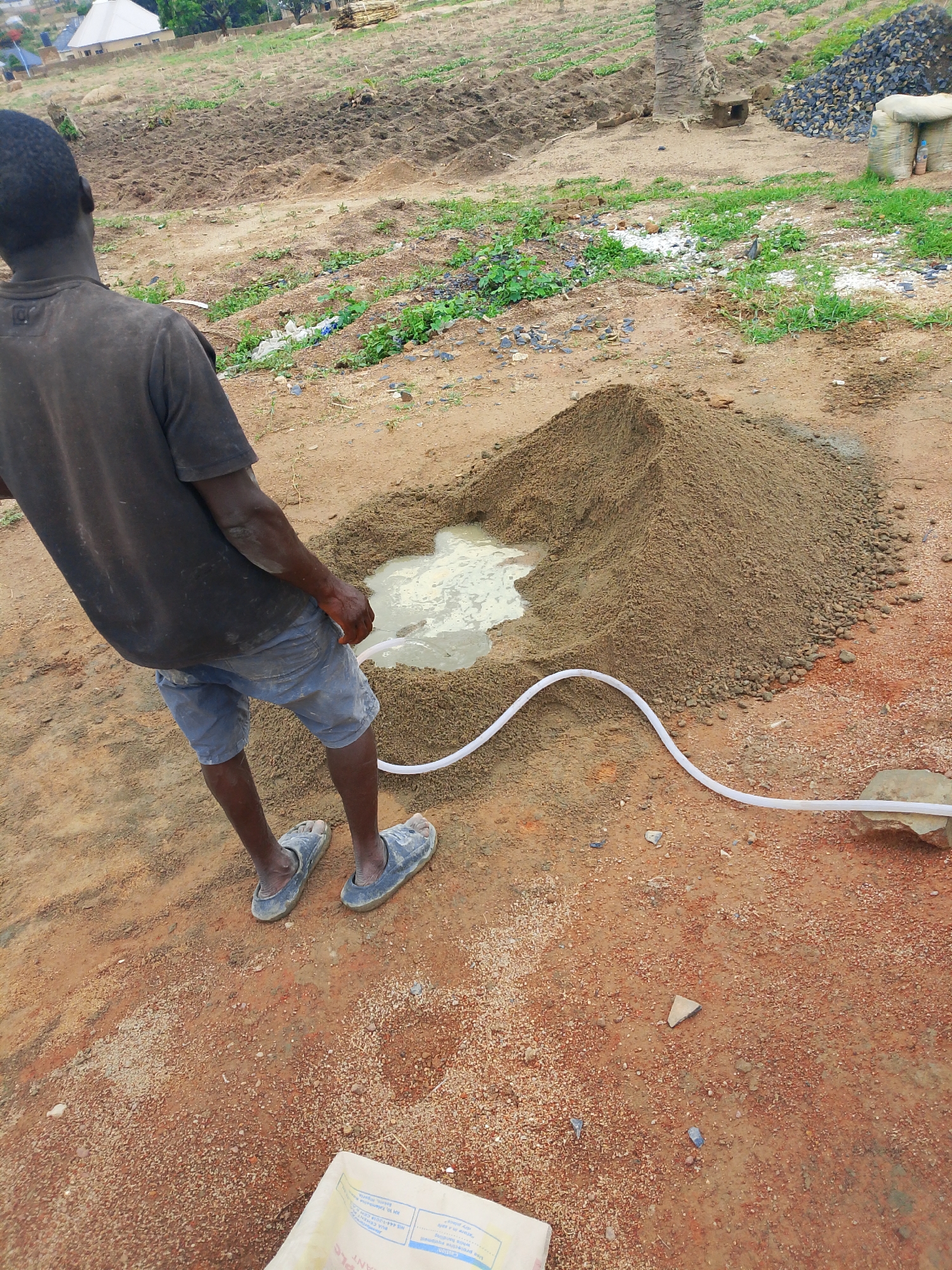 A man is mixing cement on bare ground at a construction site in Jos, Nigeria. He is standing near a mound of sand, pouring water into a crater at the top. A white plastic hose runs from off-frame to the water. The man is Black, wearing a dark gray t-shirt, light blue shorts, and blue rubber sandals. The ground is a mix of reddish-brown soil and light-colored sand. In the background, tilled land is visible, along with some low-lying vegetation and a few buildings in the distance. There is a stack of lumber in the background. A partially visible bag of cement is in the lower left corner.