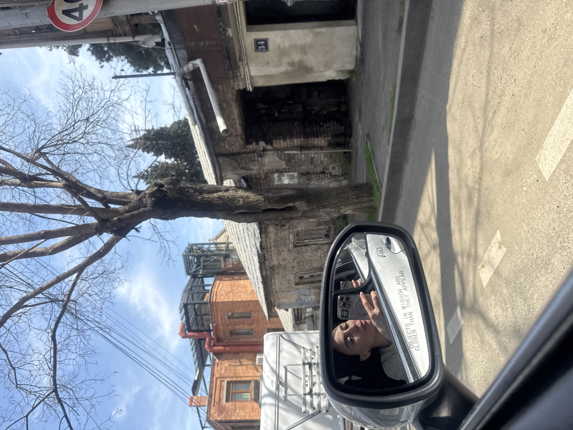 The image, taken from inside a car, shows a street scene with buildings and infrastructure in Tbilisi, Georgia. A woman is reflected in the side mirror, holding a smartphone. The mirror reads "OBJECTS IN MIRROR ARE CLOSER THAN THEY APPEAR." Outside, there's a speed limit sign with "40" on it, indicating a 40 km/h speed limit. A building with the number "34" is visible. A bare tree stands to the left of the frame against a partly cloudy sky. Other buildings with brick and metal structures are also visible, alongside power lines. The road shows lane markings.