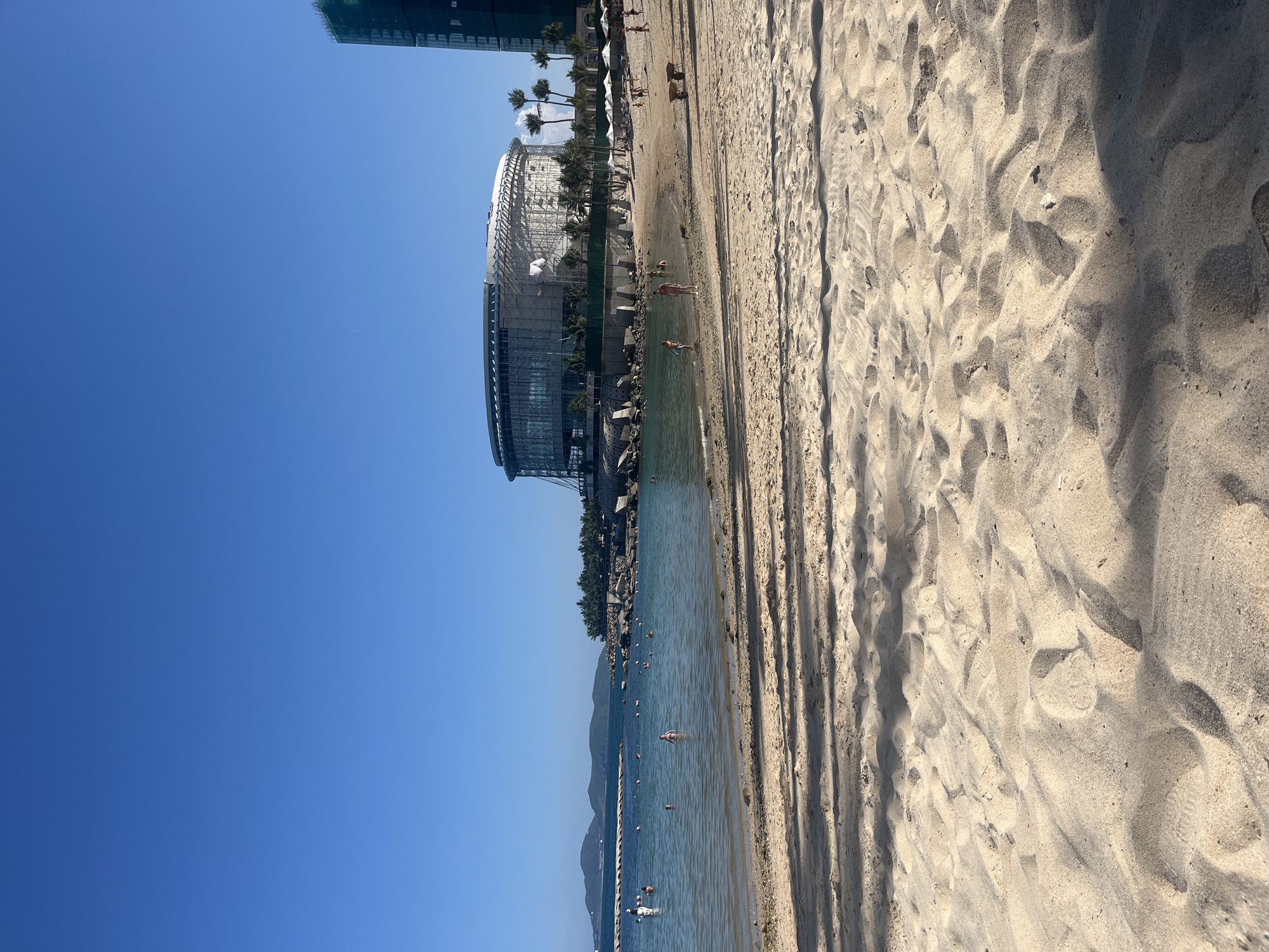 A wide shot captures a sandy beach on a clear, sunny day, with the ocean and a cityscape in the background. The foreground is dominated by textured sand, with dappled shadows suggesting foliage overhead. A gentle surf laps at the shore, and several people are visible in the shallow water, some swimming and others wading.
The midground features a crescent-shaped beach with a few scattered lounge chairs and umbrellas. Beyond the beach, a large, circular building under construction, wrapped in scaffolding, stands prominently. To its right, a modern high-rise building, partially covered in green netting, reaches towards the sky. The coastline is dotted with palm trees, and a breakwater extends into the sea. In the distance, a chain of islands forms a hazy silhouette against the horizon. The overall scene is one of a vibrant seaside resort area on a bright, clear day.