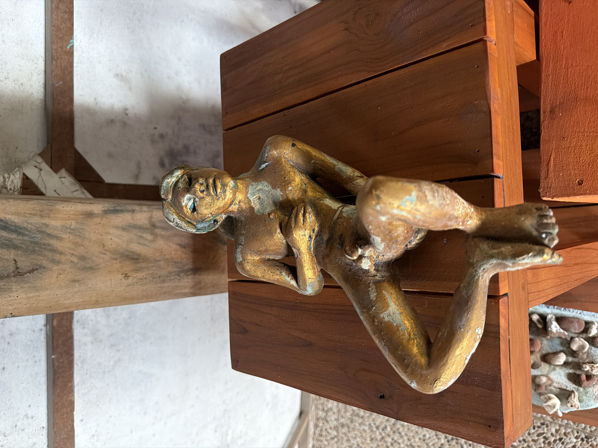 A golden statue of a reclining figure with blue eyes is positioned on a wooden surface in Tejakula, Indonesia. The statue has an aged patina with green and blue accents and is depicted in a relaxed pose, with one hand resting on its chest. The wooden surface appears to be part of a table or platform, with visible grain and planks. In the background, rough-textured walls and wooden beams are present, suggesting an indoor or covered outdoor setting. Below the table, a decorative element containing small stones and shells is partially visible. The overall lighting is subdued, casting soft shadows on the statue and its surroundings.