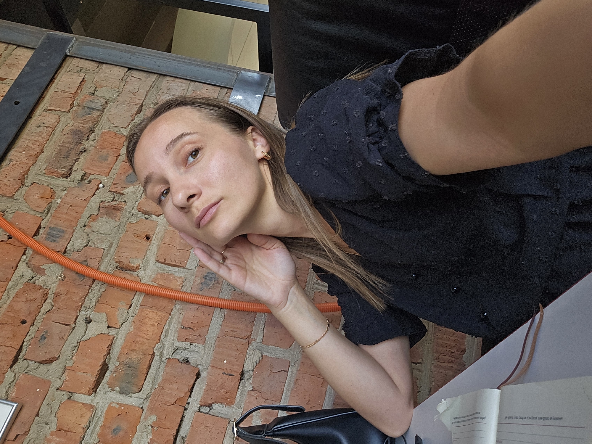 A young woman is angled toward the camera in an indoor location that appears to be in the city of Chaikovsky, Russia. The background reveals a brick wall with an orange conduit running along it, along with a metal structural element overhead. The woman has light brown hair and is wearing a black top. She is resting her cheek on her hand. A bracelet adorns her wrist. A black purse sits beside her. A book and brown leather straps are visible on a white surface, suggesting a table or desk.
