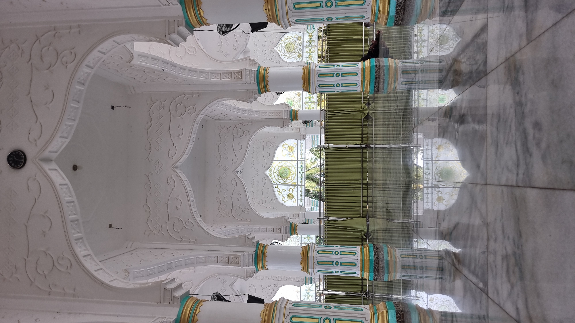 The image depicts the interior of a building with Islamic architectural elements. White columns with gold and teal accents support arched ceilings decorated with intricate relief patterns. Green curtains hang behind metal railings, and beyond them, arched windows with decorative glass are visible, offering a glimpse of trees and sky. The highly polished marble floor reflects the columns, arches, and windows, creating a symmetrical visual effect. In the lower right corner, a person is lying down on the floor. A black clock is mounted on the ceiling in the upper center of the image. The overall scene suggests a serene and ornate interior space, likely a mosque or a similarly significant building.