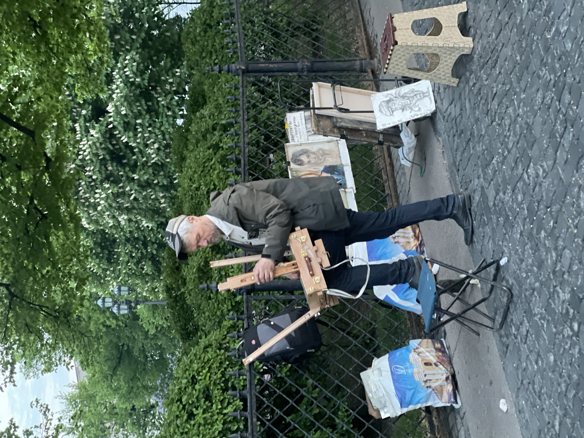 An older man is setting up an easel on a sidewalk in what appears to be Saint Petersburg, Russia. He is wearing a green jacket, dark pants, black shoes, and a cap. A black metal fence is behind him, with greenery visible beyond the fence. Various artwork pieces are leaning against the fence, along with a folding step stool. The ground is paved. Bags with images of what looks like a building in Saint Petersburg are nearby. He is standing next to a folding chair.