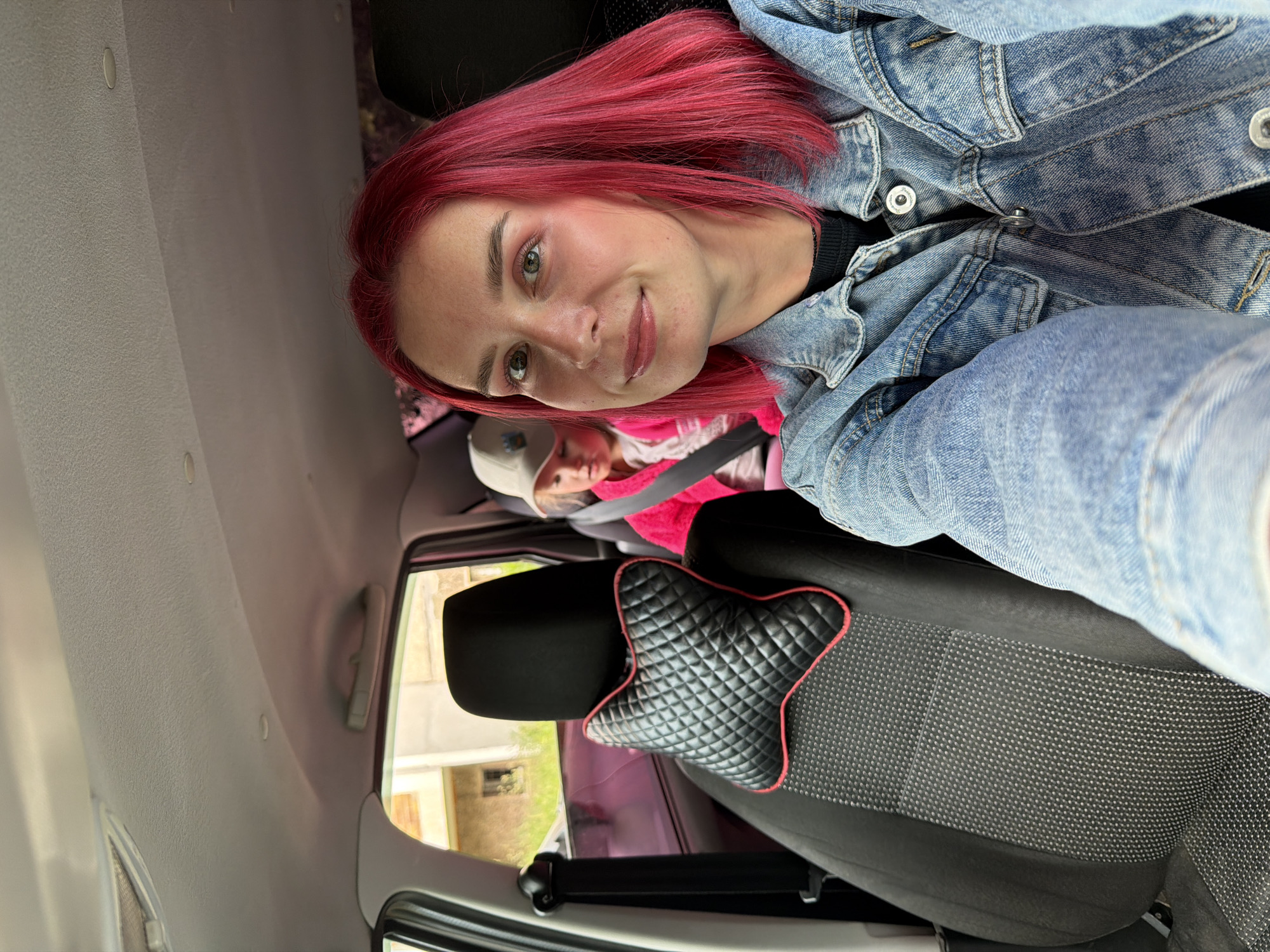 A tilted selfie captures a young woman and a child inside a car, likely in Chaikovsky, Russia. The primary subject, a woman with striking bright pink/red hair, light skin, and green eyes, smiles faintly at the camera. She is wearing a light blue denim jacket over a dark shirt.
In the background, a child is visible, wearing a light-colored baseball cap and a pink top, seemingly secured in a car seat or booster seat. The car's interior features light gray textured ceiling material, dark gray fabric seats, and a distinctive black, star-shaped pillow with pink trim on a headrest. Through the car windows, a partial view of a building with light walls and some foliage suggests an outdoor, possibly urban or suburban environment.
The image appears to be taken during the daytime under diffused natural light. The activity is a casual selfie, capturing a moment inside a vehicle. No clearly legible text is visible in the frame.