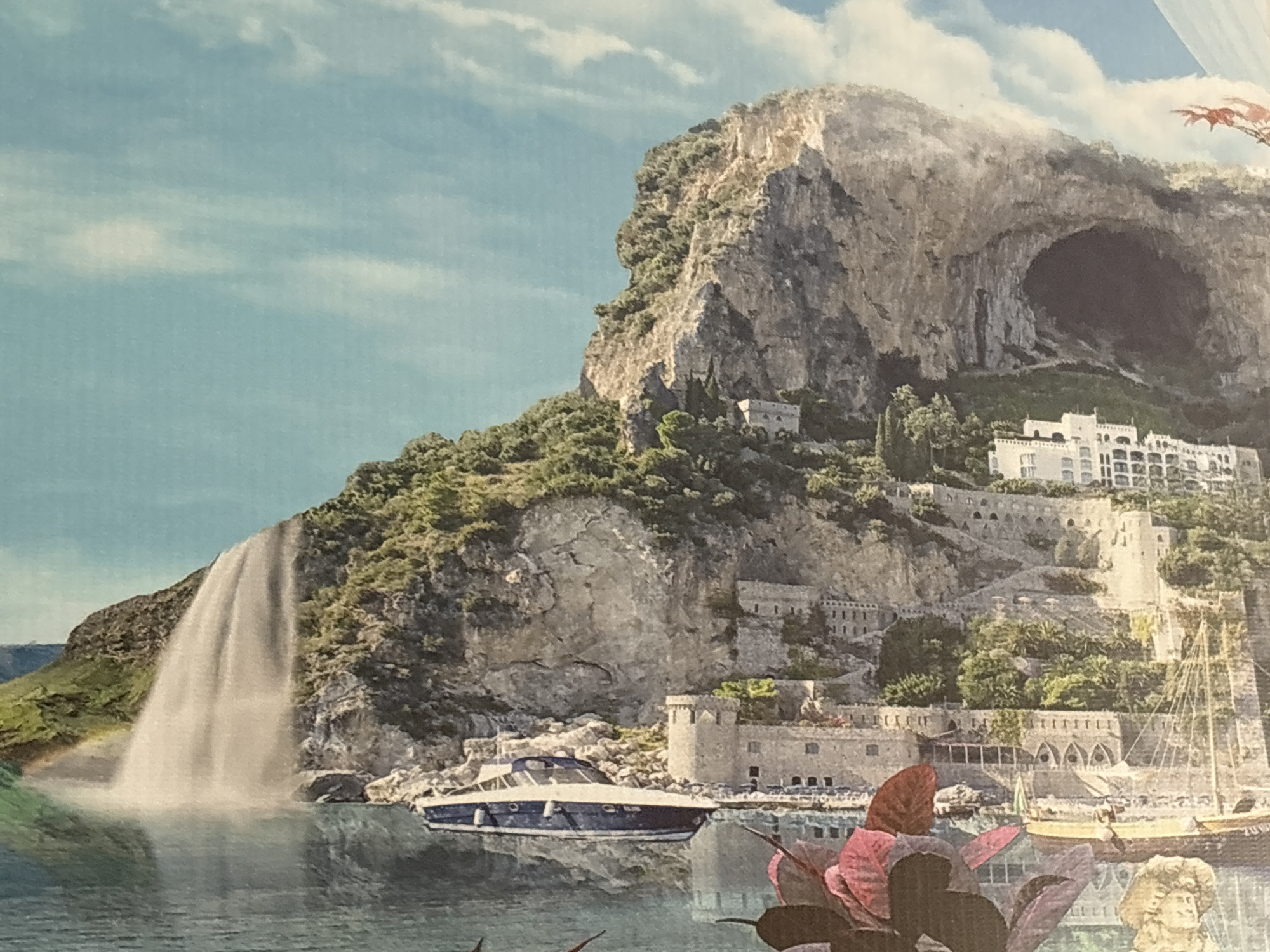 This media file captures a vibrant, idealized coastal landscape, resembling a digital collage or an intricate painting. The scene is dominated by a majestic, rugged cliff face on the right, covered with lush green foliage and featuring a large, dark cave entrance high up. Below the cliff, a series of white, terraced buildings, possibly a town or resort, are nestled into the hillside, showcasing traditional architecture with arches and multiple levels.
To the left, a powerful waterfall cascades into a body of calm, reflective water, which could be the sea or a large lake. A faint rainbow arch is visible at the base of the waterfall. In the water, a sleek blue and white modern yacht is prominent in the mid-ground, while a classic sailboat with intricate rigging is partially visible further right, appearing docked near the coastal structures. Some buildings closer to the water have a fortress-like appearance with towers and fortified walls.
The sky is a bright, clear blue with scattered white clouds, suggesting a sunny day. In the very bottom right foreground, parts of a large, dark-leaved plant with reddish-pink petals or flowers are cut into the frame, along with a small, ornate, light-colored statue or carving, possibly a cherub. The overall impression is one of a dramatic and picturesque Mediterranean-style setting, possibly a fantasy interpretation or a composite of various beautiful elements. There is no visible text in the image.