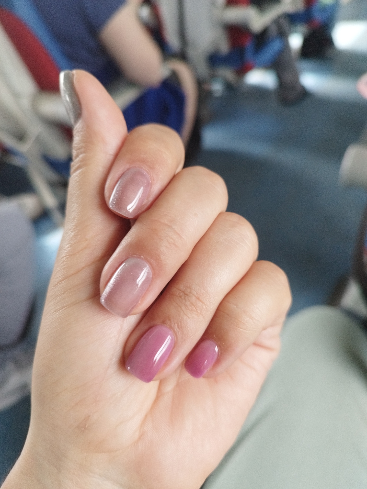 A close-up, eye-level, outdoor shot focuses on a person's left hand, displaying manicured nails. The nails are coated in a sheer, shimmery nude polish on the thumb, index, and middle fingers, and a solid, muted pink polish on the ring and pinky fingers. The background is out of focus, showing blurry figures in casual attire, suggesting a public or semi-public space like a park or transportation. There is no visible information to determine the location in Saint Petersburg, Russia.