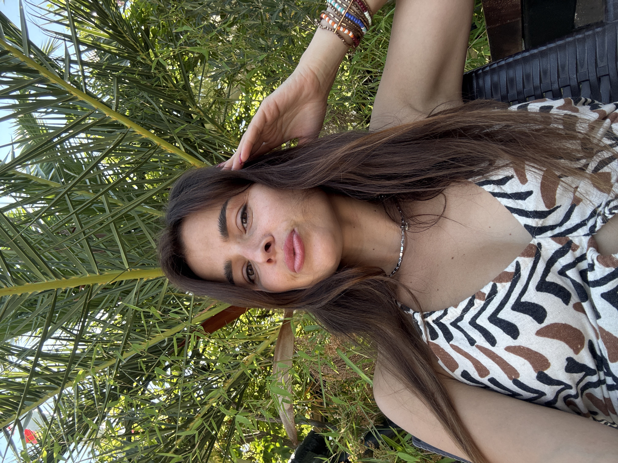 This media file is a portrait of a young woman taken outdoors, possibly on a sunny day given the bright lighting and the presence of lush greenery. She is lying down, with her head propped up by her hand, and her gaze is directed towards the camera. The woman has long, dark brown hair that falls around her shoulders, and she is wearing a patterned top with black, brown, and white markings that resemble animal print. A beaded bracelet adorns her wrist, and she wears a silver necklace.
The background is filled with the fronds of a palm tree and other foliage, suggesting a tropical or subtropical location. Sunlight filters through the leaves, creating dappled light. The woman's expression is relaxed and perhaps a little playful. The photo appears to be a selfie, given the close-up perspective and the angle of the shot. There is no visible text in the media file.