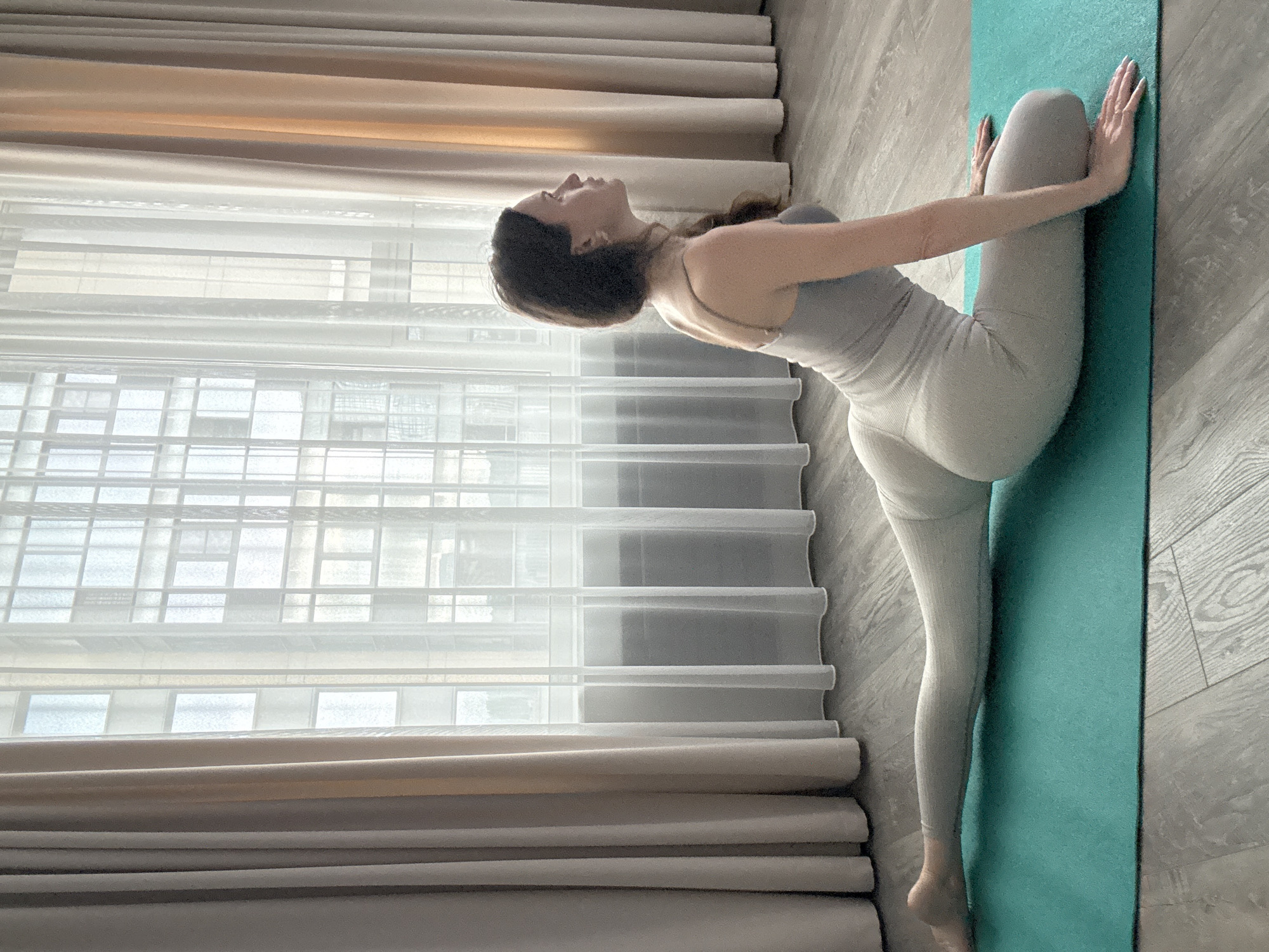 A woman is performing a yoga pose on a teal mat in a room with a large window and floor-to-ceiling curtains. She is wearing a light grey athletic outfit. Her right leg is extended behind her, and her left leg is bent at the knee with her foot tucked under her. Her hands are placed on the mat in front of her. The room appears to be a home environment, possibly an apartment in Khimki, Russia, given the style of the window and curtains. The floor is made of light-colored wood. The curtains are light beige and white. The lighting suggests it is daytime.