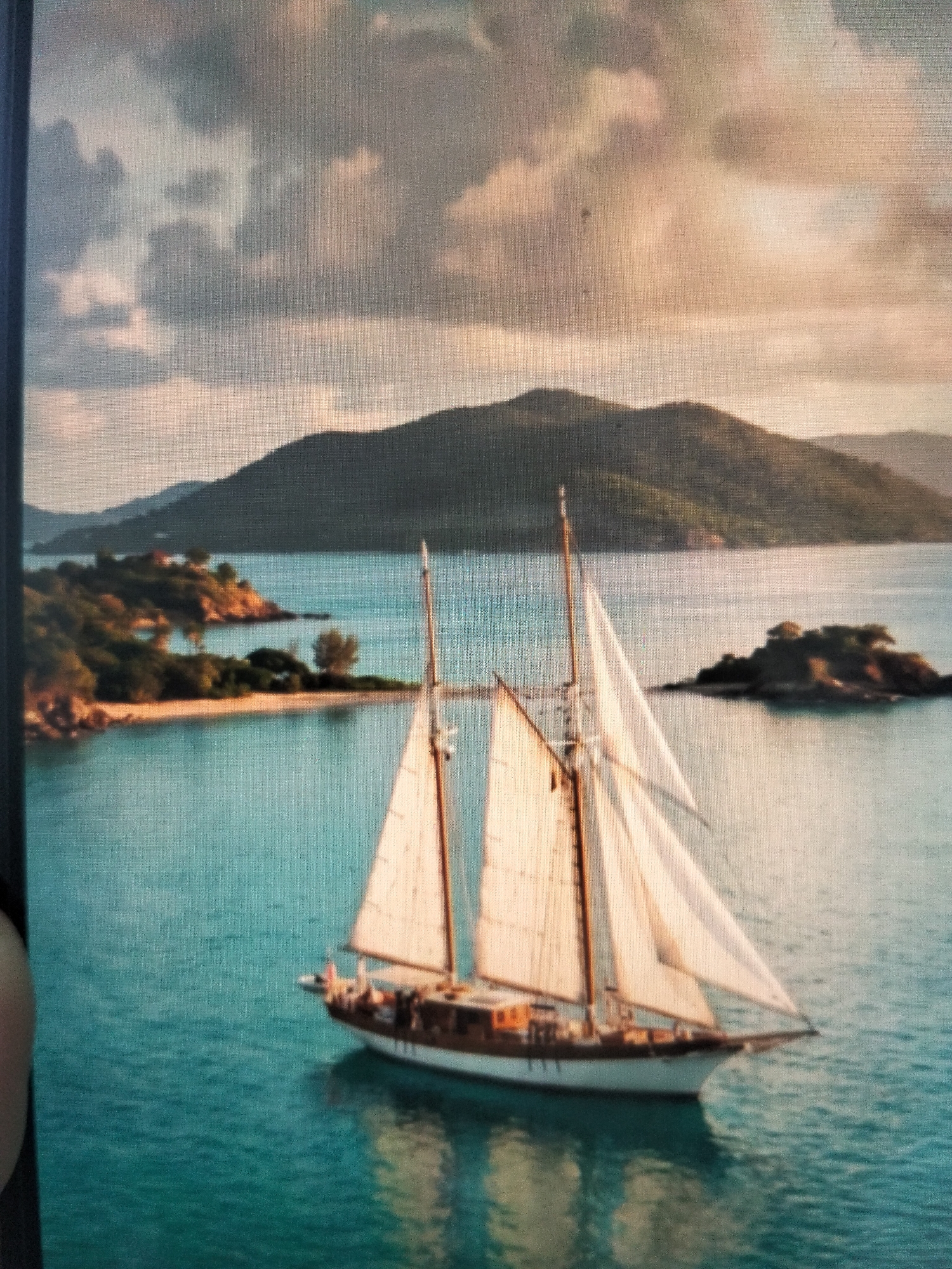 An aerial shot shows a schooner sailing on turquoise water near a tropical island. The schooner has two masts and white sails. The island is covered in green trees and has a small sandy beach. In the background, more islands and hills are visible under a cloudy sky. The overall scene suggests a peaceful, idyllic setting. There are no visible inhabitants or buildings in the city of Payagaboh, Indonesia.