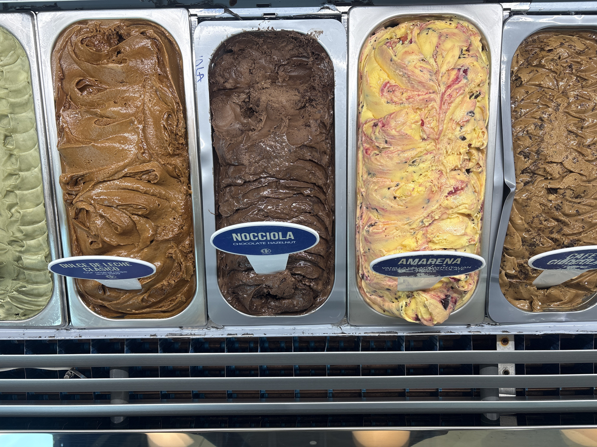 The image displays a close-up, top-down view of five rectangular stainless steel bins containing various flavors of gelato or ice cream, likely within a commercial display freezer in Huixquilucan de Degollado, Mexico.
From top to bottom, the visible contents and their associated labels are:
1. A partially visible bin containing a light green, smooth substance.
2. A full bin of textured, medium-brown gelato labeled "DULCE DE LECHE CLÁSICO" (Classic Dulce de Leche) on a blue and white oval marker.
3. A full bin of dark brown, textured gelato labeled "NOCCIOLA CHOCOLATE HAZELNUT" (Chocolate Hazelnut), also with "CHOCOLATE Y AVELLANA" and an "AK" logo.
4. A full bin of yellow gelato with prominent red swirls and dark specks, labeled "AMARENA VANILLA GELATO WITH ITALIAN CHERRIES," also with "GELATO DE VAINILLA CON CEREZAS ITALIANAS."
5. A partially visible bin of light brown gelato with dark granular inclusions, with a blue and white label partially visible, indicating "CAFE" (Coffee).
The gelato surfaces exhibit peaks and troughs consistent with being recently scooped or prepared. To the left of the bins, a vertical metallic structure with horizontal silver bars and reflective surfaces is visible, suggesting the side of the display unit. No individuals, actions, or specific interactions are depicted. The environment appears to be an indoor commercial setting, specifically a gelato shop or a similar establishment.