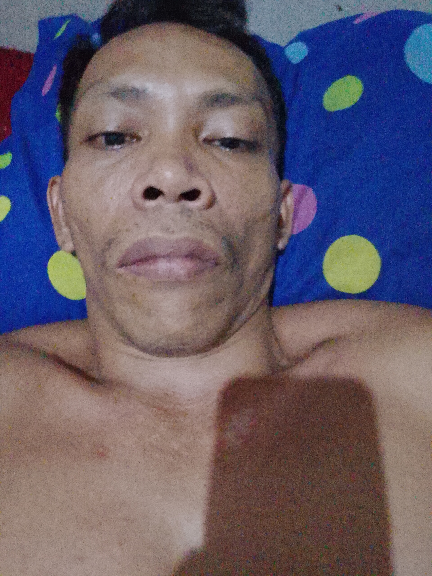 A shirtless man is lying down with his head on a blue pillow with yellow, pink, and blue polka dots. He is facing the camera. The location is Bukit Ketok, Indonesia.