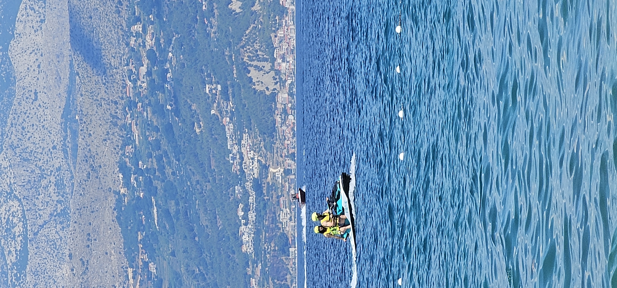 Two individuals, wearing yellow life vests and helmets, are riding a blue and white jet ski on a clear, blue body of water. The jet ski is creating a white wake behind it. In the background, across the water, a coastal city with white buildings and red rooftops is nestled at the base of a lush, green mountainside. The mountains in the distance are rocky and sparsely vegetated, with a clear blue sky overhead. The scene suggests a recreational activity in a scenic, coastal location, likely during daylight hours. The location is the city of Supetar, Croatia, on the island of Brač.