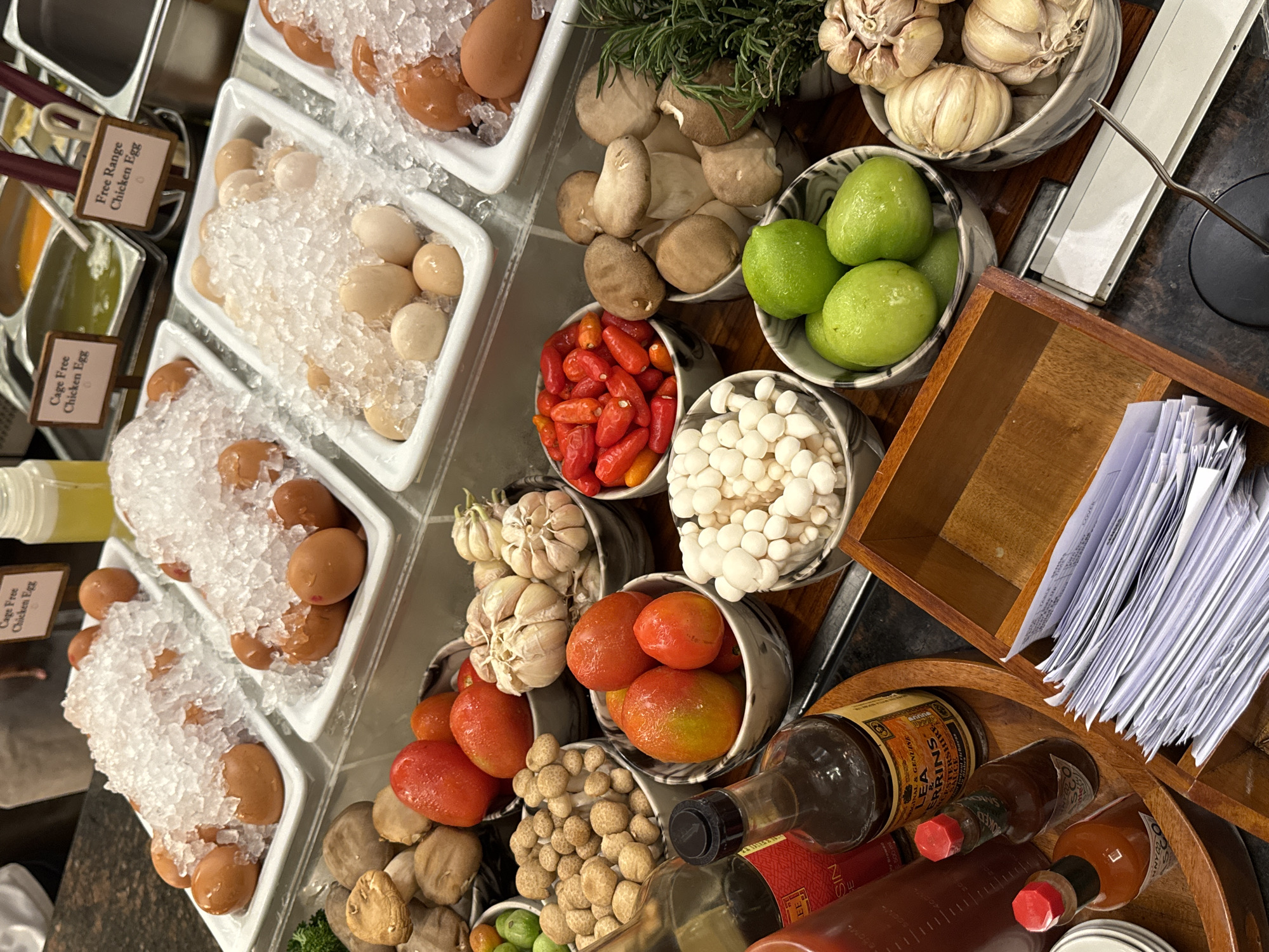The image depicts a buffet station with a variety of fresh ingredients displayed in bowls and containers. In the background, there are two white rectangular trays filled with eggs, some on ice. Signs above the eggs indicate "Cage Free Chicken Egg" and "Free Range Chicken Egg." In front of the eggs, various vegetables and mushrooms are arranged. There are bowls containing tomatoes, garlic bulbs, small red chili peppers, small white mushrooms, and green limes. Several bottles of sauces, including what appears to be Lea & Perrins Worcestershire Sauce and Tabasco sauce, are placed on a wooden tray. A wooden box filled with stacks of white paper is also visible. The overall environment suggests a food service setting, possibly a restaurant or hotel buffet in Kuta Selatan, Indonesia.