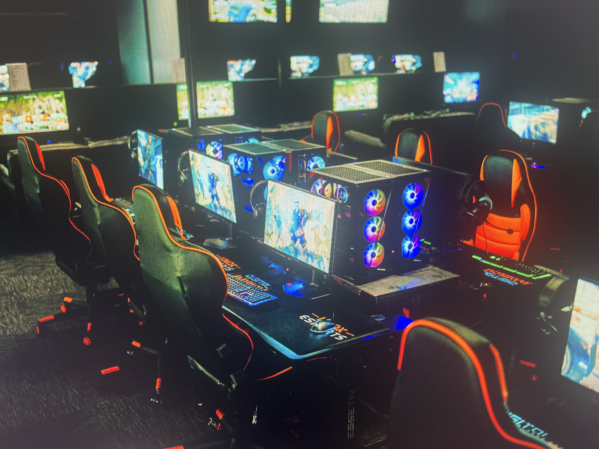 The image displays the interior of a modern esports arena or gaming café, possibly located in Varna, Bulgaria. The scene is dimly lit, creating an immersive atmosphere, with most illumination originating from the numerous computer screens and the vibrant RGB lighting of the gaming hardware.
The facility features multiple rows of high-performance gaming stations. Each station is equipped with a black gaming desk, an ergonomic gaming chair distinguished by black upholstery and striking orange accents, a large monitor displaying colorful game graphics, and a powerful gaming PC. The PC towers have transparent side panels, revealing internal components and multiple RGB-lit fans that glow in shades of blue, purple, and other colors. Each desk is also outfitted with an RGB-backlit keyboard, a mouse, and headphones, indicating readiness for immediate use.
Additional monitors are visible mounted on the walls above the gaming stations, displaying various graphical content, likely game streams, scores, or promotional material. No people are present in the image, suggesting a time outside of peak hours, such as early morning or late evening. The overall environment conveys a high-tech, professional, and inviting setting designed for competitive or casual gaming.
Visible text includes "INDX ESPORTS" and "ESSETIV" (or "ESSETV") printed on the surface of the gaming desks or mousepads. Partial text "DIGITAL" can also be observed on one of the gaming chairs.
