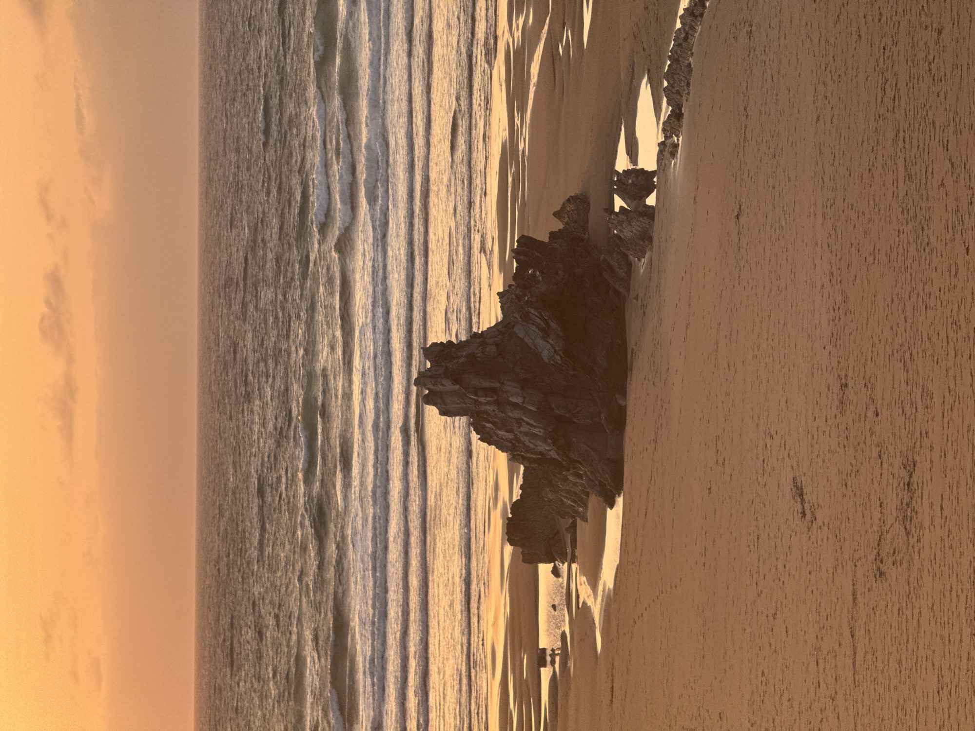 The image depicts a coastal scene at sunset in Sopela, Spain. The sky is a warm, orange hue, transitioning into a soft pink, with scattered clouds. The ocean stretches to the horizon, with gentle waves breaking on the shore. In the foreground, a sandy beach is marked by tidal pools reflecting the sunset light. A prominent rock formation, dark and jagged, juts out from the sand, with smaller rocky outcrops scattered around it. There are no people or animals visible in the immediate vicinity of the rock formation. The overall impression is one of a tranquil, natural landscape bathed in the warm light of dusk.