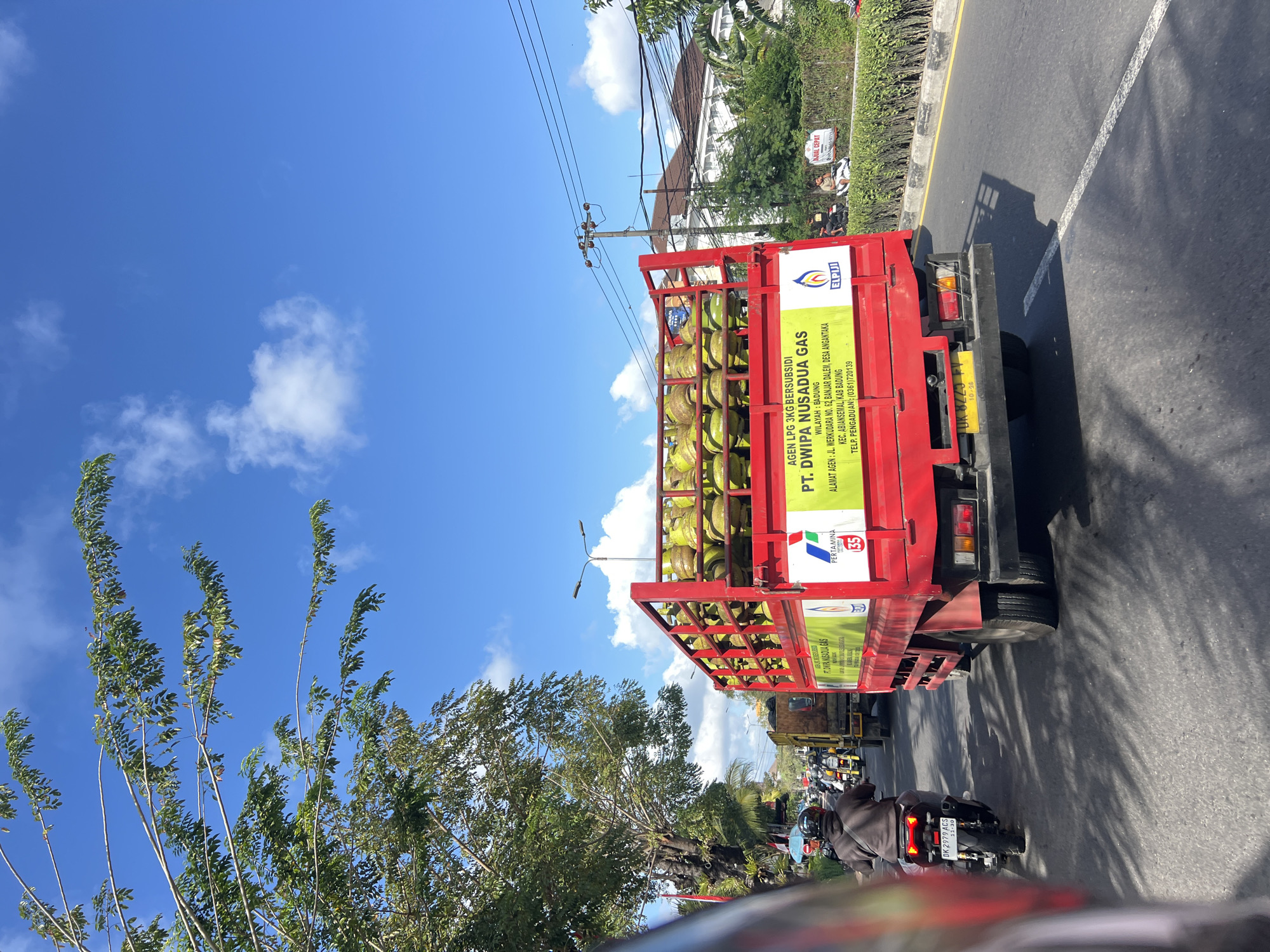 The image depicts a street scene in Kuta, Indonesia, on a sunny day with a clear blue sky and scattered clouds. A red truck, laden with numerous yellow propane gas cylinders, is the primary subject. The truck has text on its side, including "AGEN LPG 3KG BERSUBSIDI PT. DWIPA NUSADUA GAS" and an address. To the left of the truck, a person on a motorcycle with the license plate "DK 2979 ACS" is visible, traveling in the same direction as the truck. Further ahead, another truck and more motorcycles are present on the road. The environment features trees lining the street and buildings in the background, with power lines overhead. The road is paved, and there are road markings and a curb with a hedge.