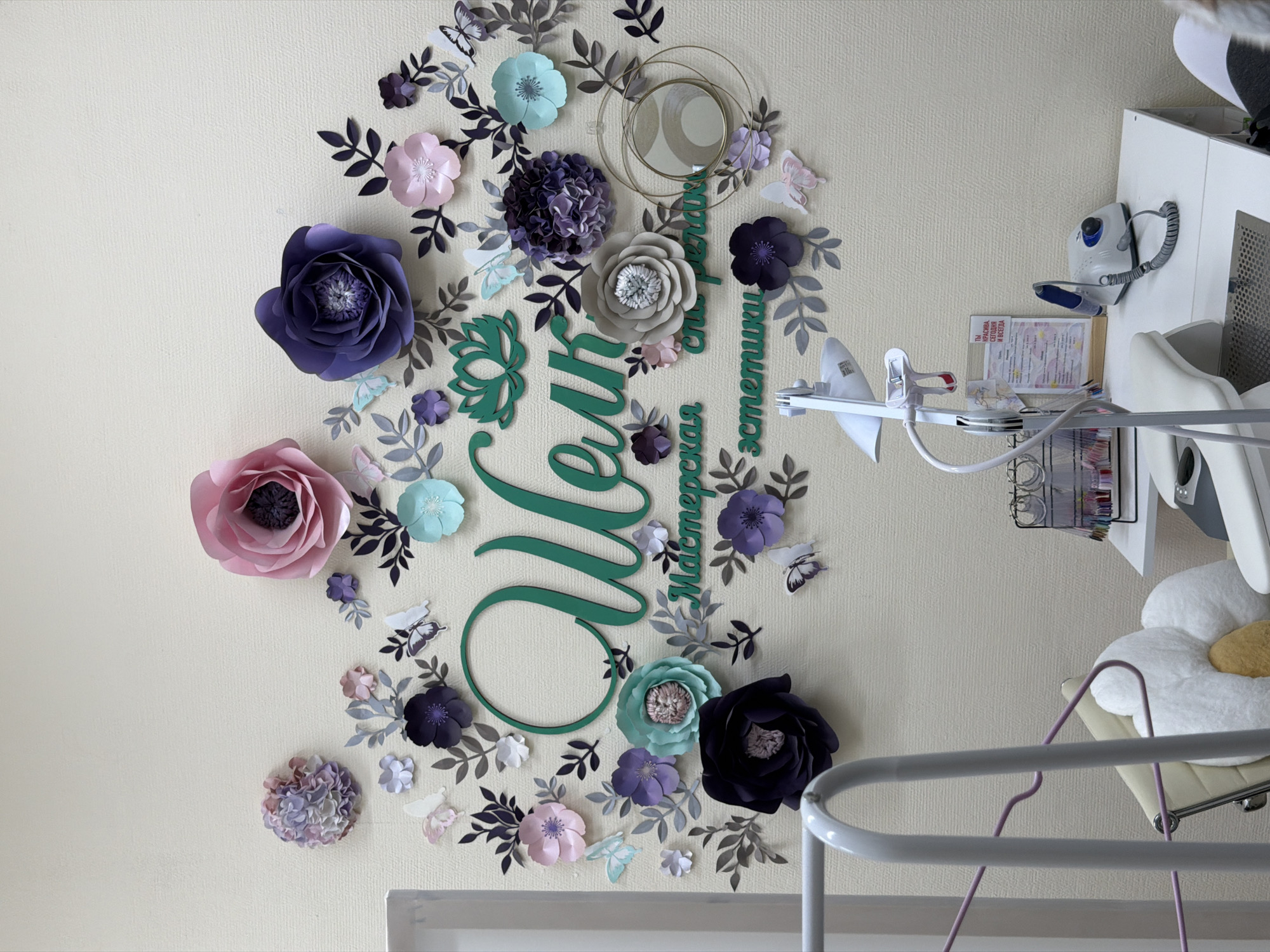 The image shows a wall decoration in what appears to be a business setting, possibly in Chaikovsky, Russia based on the Cyrillic text incorporated into the design. The decoration features oversized paper flowers in shades of purple, pink, teal, and gray arranged around a cursive script spelling "Olga, Aesthetic Studio" (translated). Small paper butterflies and leafy cutouts are scattered around the flowers and text.
The wall is a light cream color with a textured surface. To the right of the floral decoration, medical equipment is visible including a surgical light, equipment mounted on a white cabinet, and a chair with a daisy pillow.