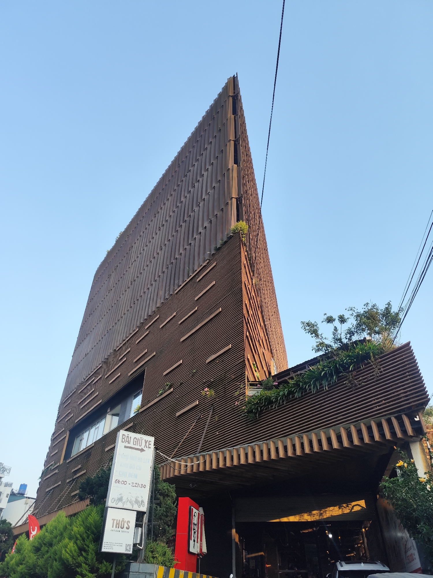 The image captures a uniquely shaped building in Da Lat, Vietnam. The building's facade is clad in brown horizontal slats, giving it a textured appearance. The design incorporates greenery, with plants and small trees visible on ledges and the roof.
A sign with Vietnamese text and graphics for "Bãi Giữ Xe" (parking) is visible near the building's base, along with a sign that says “THO’S ARTISAN COFFEE." A red KFC sign is present, and other signage indicates a commercial presence.
The structure has a triangular or wedge-like shape that narrows towards the top. Power lines run across the clear blue sky above. Street-level elements, including glimpses of vehicles and greenery, suggest a street scene. The perspective of the shot is looking up at the building.