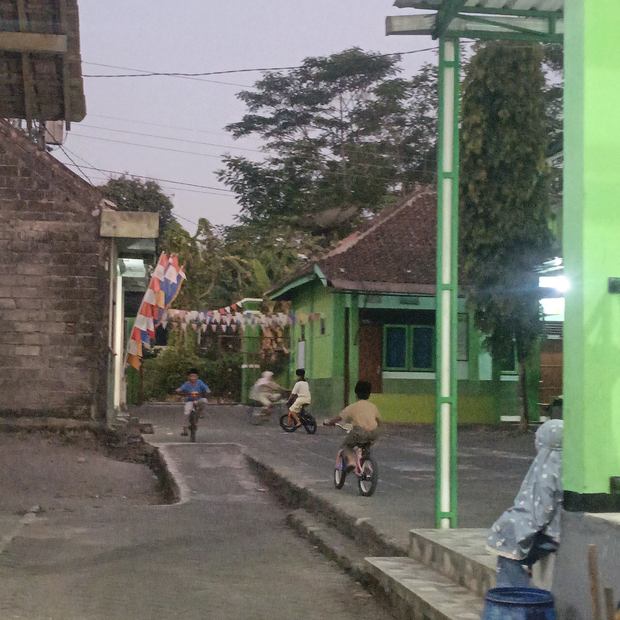 In an outdoor scene in Salatiga, Indonesia, on a paved street, a group of children are riding bicycles in what appears to be a residential area. The sky is overcast, suggesting early morning or late afternoon. On the left side of the image, a weathered brick building with a wooden overhang is visible, with electrical wires running overhead. To the right, a vibrant green building with a brown tiled roof stands, decorated with colorful triangular flags strung across its front. Several children are engaged in cycling: one child in a blue shirt rides towards the foreground on the left, while another in a white shirt is in the middle distance on a smaller bicycle. Further to the right, a child in a light brown shirt is also on a bicycle. The background features lush green trees and vegetation, common in Indonesian landscapes. A person wearing a headscarf and patterned clothing is partially visible on steps on the far right, with a blue barrel nearby. The overall impression is of a quiet, local street life, with children enjoying recreational activity.