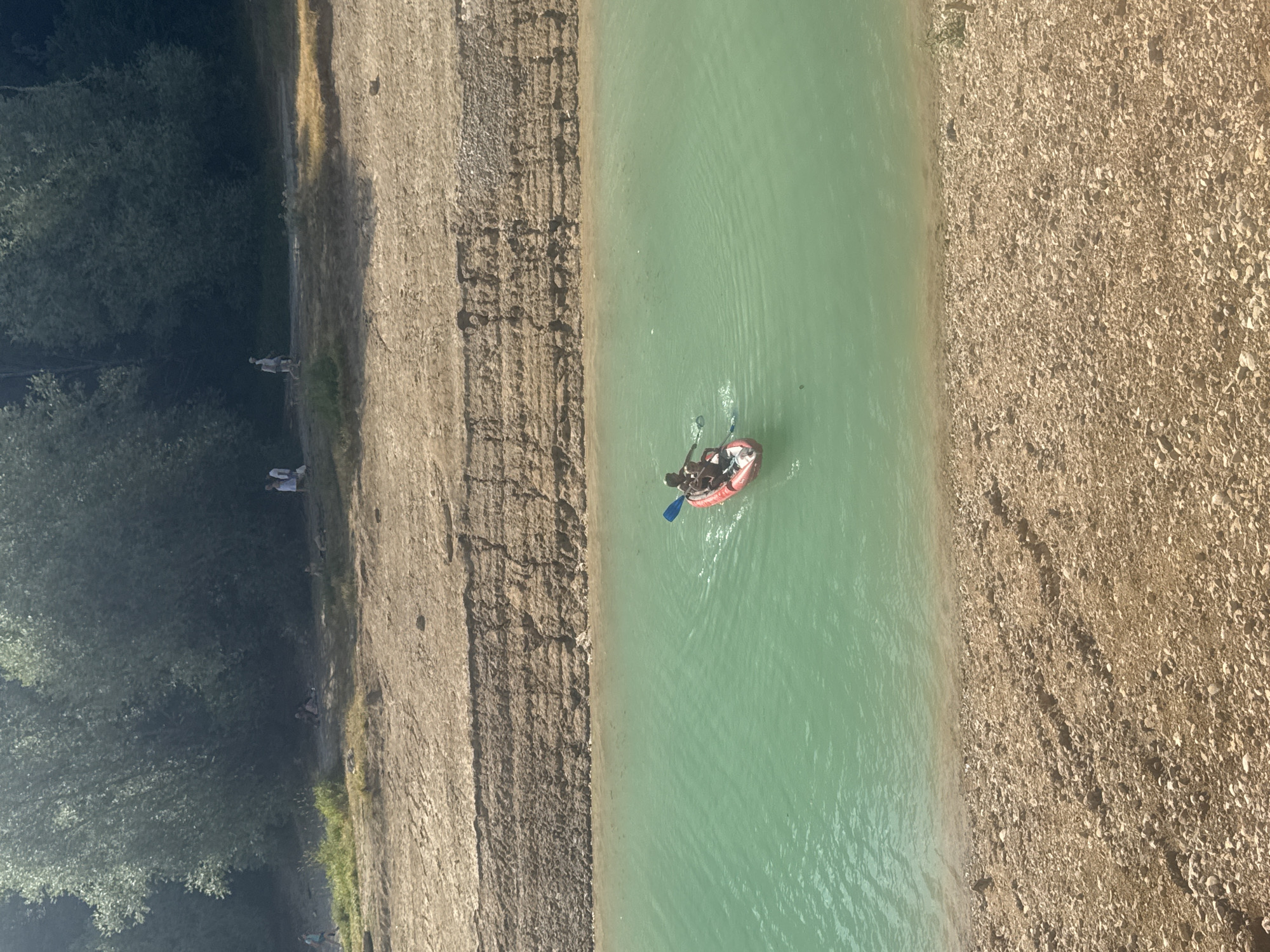 In the city of Maisod, France, a kayak carrying two individuals navigates the aquamarine waters of a lake. The kayak, a vibrant red, is positioned in the center of the body of water, with the occupants actively paddling. The surrounding environment is characterized by a rugged, sandy shoreline that transitions into a dense forest of green trees at the upper edge of the frame. In the distance, on the shoreline, several other individuals are visible, some walking and others resting. The scene depicts a recreational day outdoors, with people enjoying the lake and its natural surroundings.