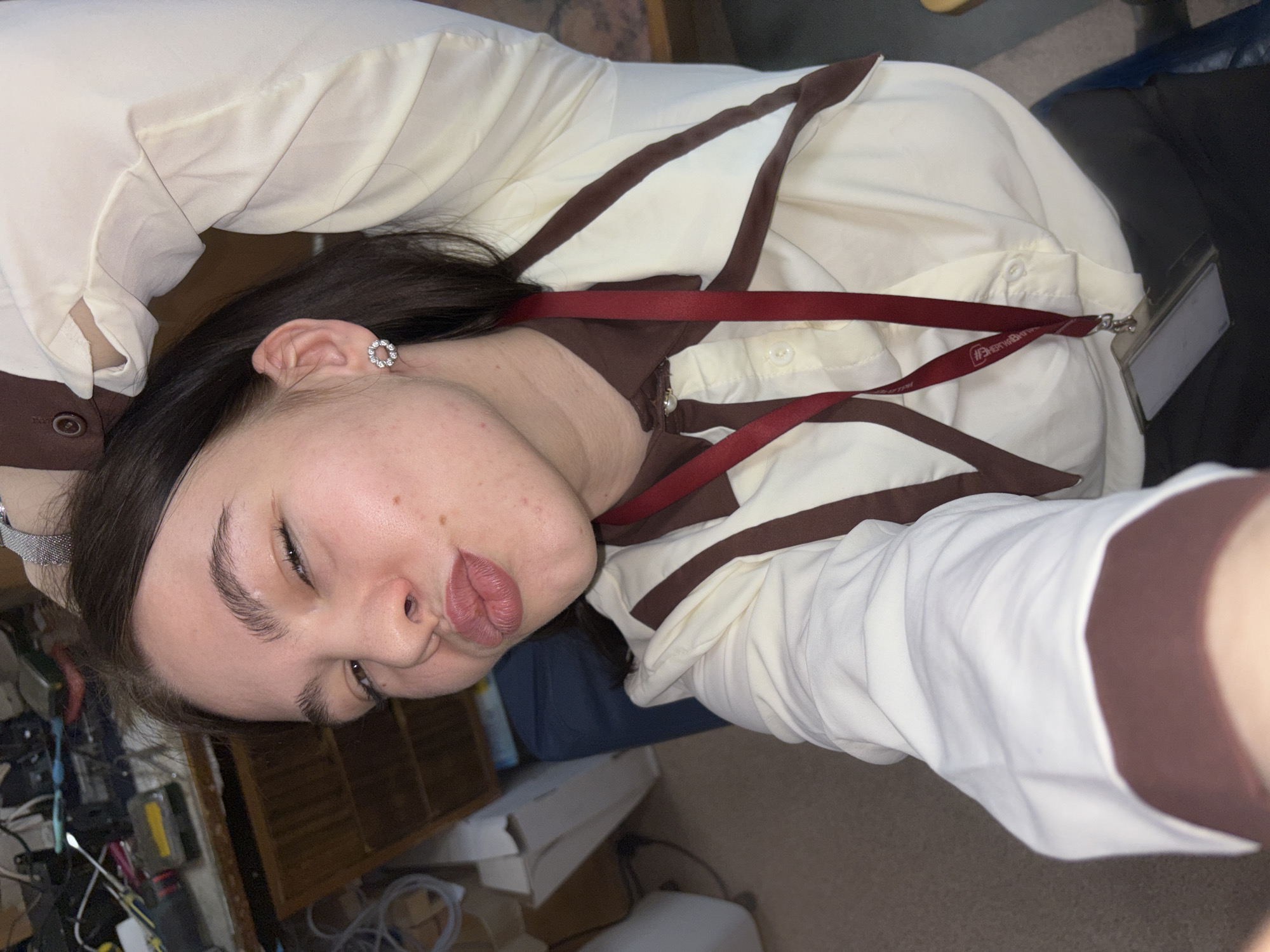 A selfie photograph shows a fair-skinned person with dark hair wearing a cream-colored button-down shirt with brown trim and a brown lanyard with a rectangular white ID card attached. The person has light brown eyes, prominent eyebrows, and is making a puckered-lip expression. A silver hoop earring is visible on their left ear. The person is positioned in front of a cluttered backdrop that includes shelves, electronic components, and various unidentified objects. The scene appears to be indoors, possibly in a private residence. There is no evidence suggesting the location of Perm, Russia.